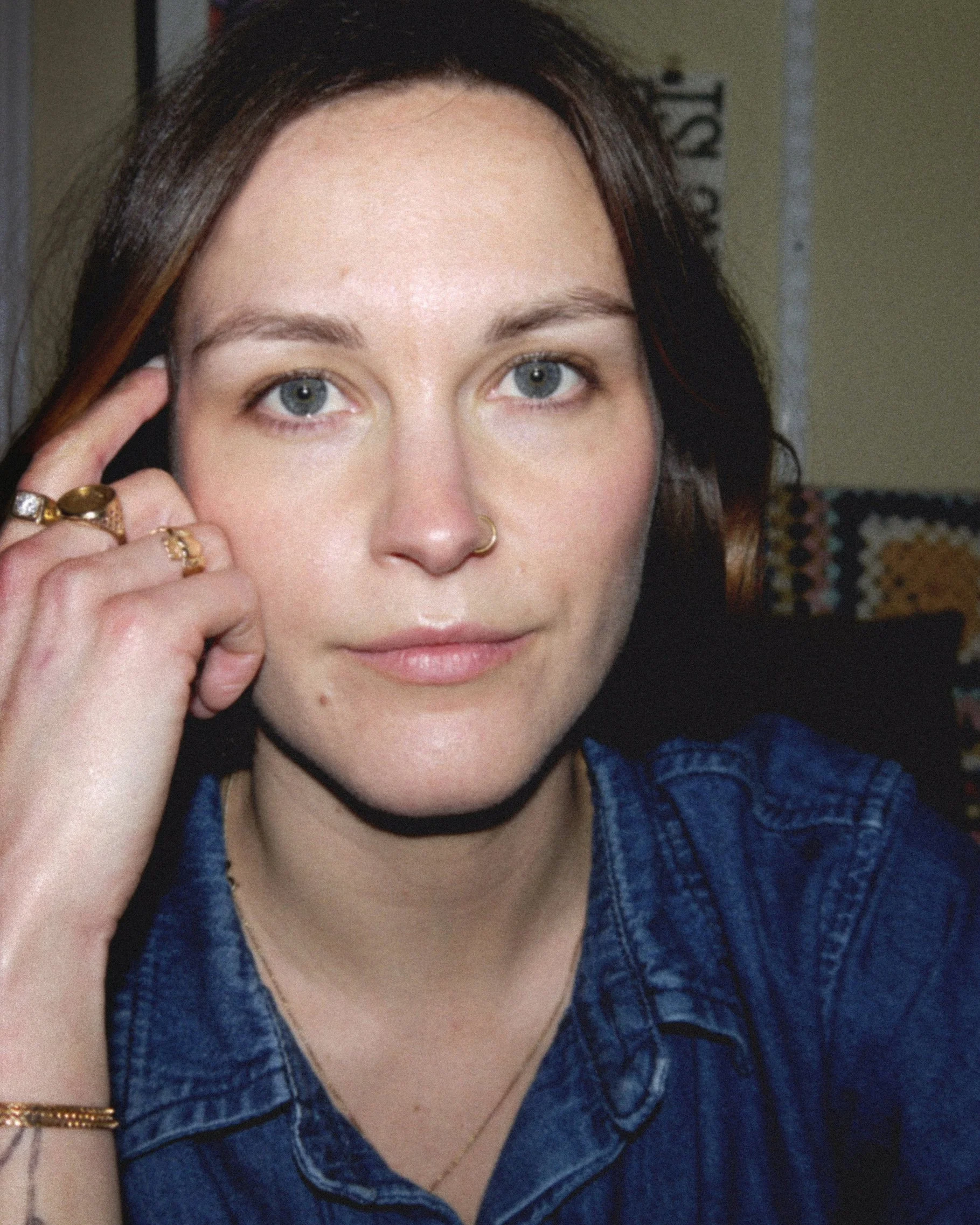 Close-up of a young woman with short dark hair, blue eyes, a nose ring, and multiple rings on her fingers, wearing a denim shirt, smiling softly.