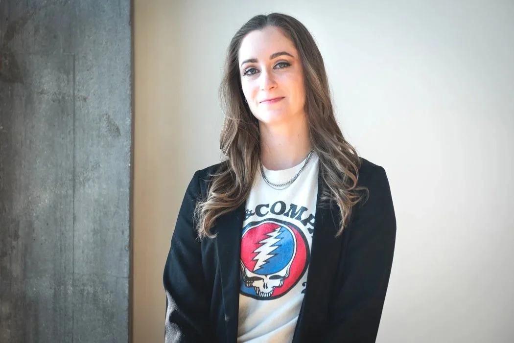 A woman with long brown hair wearing a white Grateful Dead t-shirt and black blazer, standing indoors against a wall.