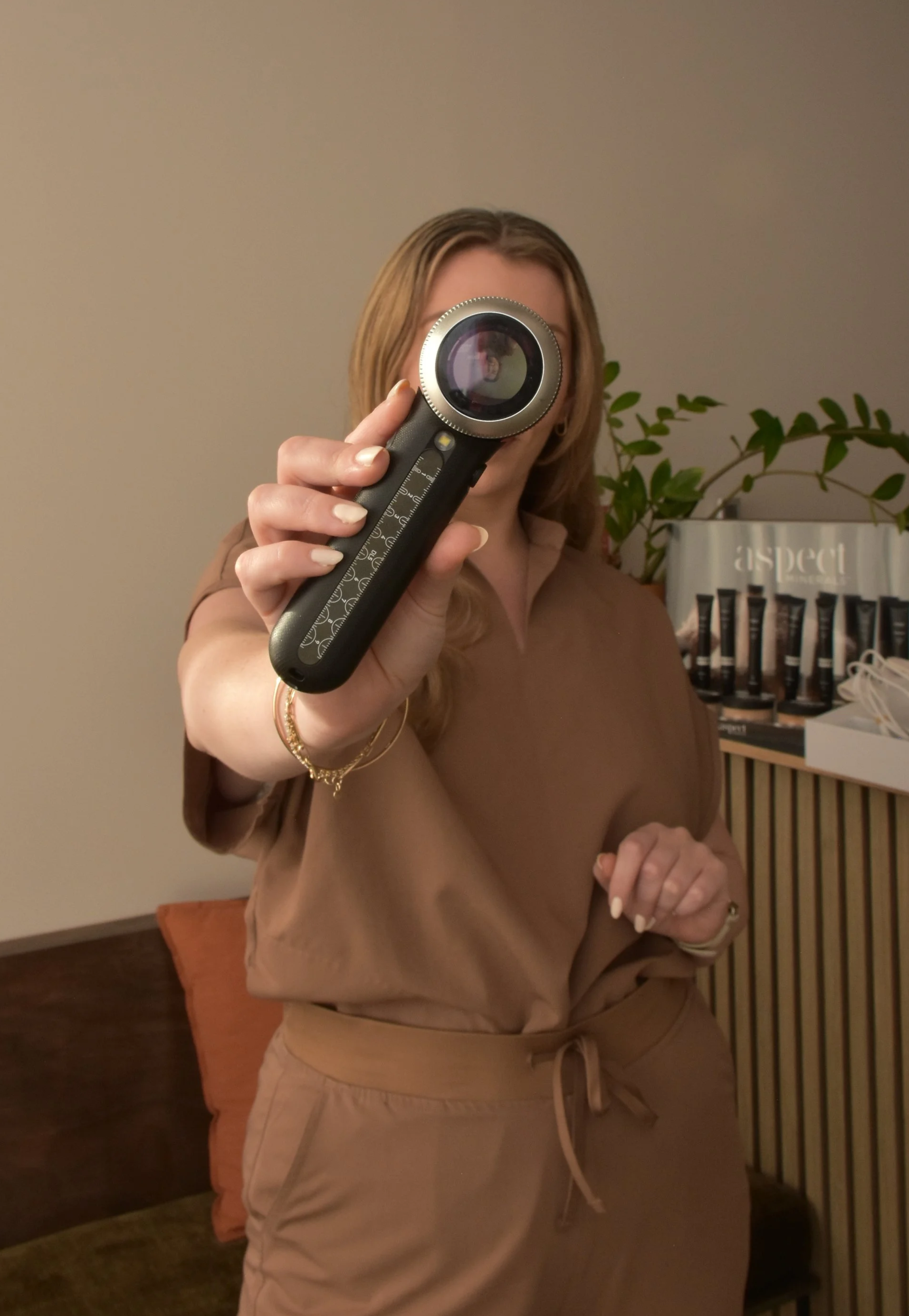 A woman (Dr. Cherie Wyatt-Hutana) with blonde hair taking a mirror selfie with a camera, wearing a brown top and beige pants, with a plant and a display of skincare products in the background.