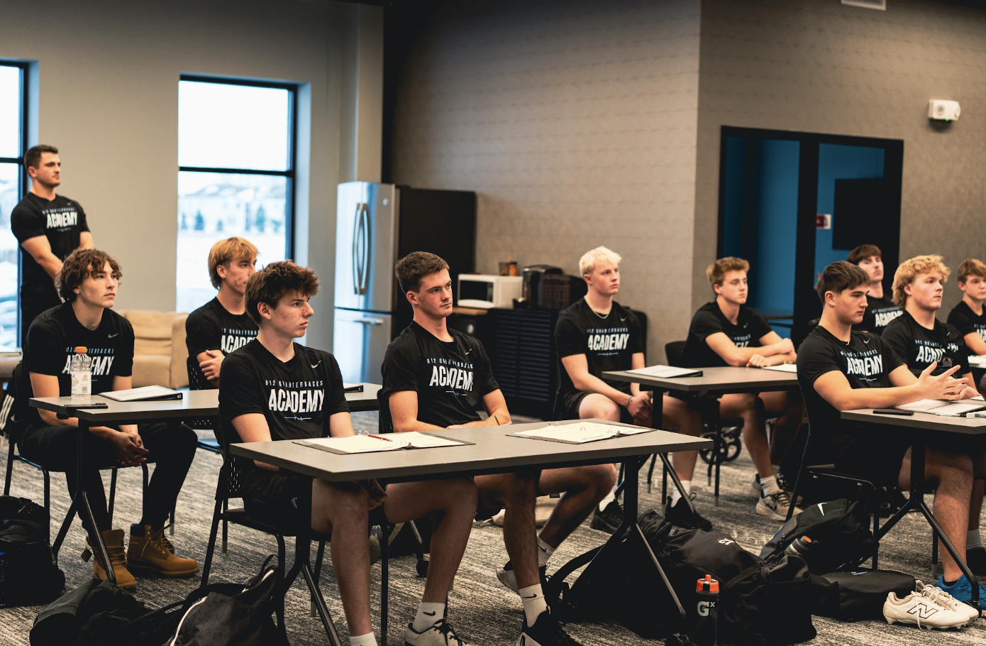 Young athletes wearing black shirts with 'ACADEMY' printed on them, sitting attentively at tables during a classroom or training session in a modern indoor facility.