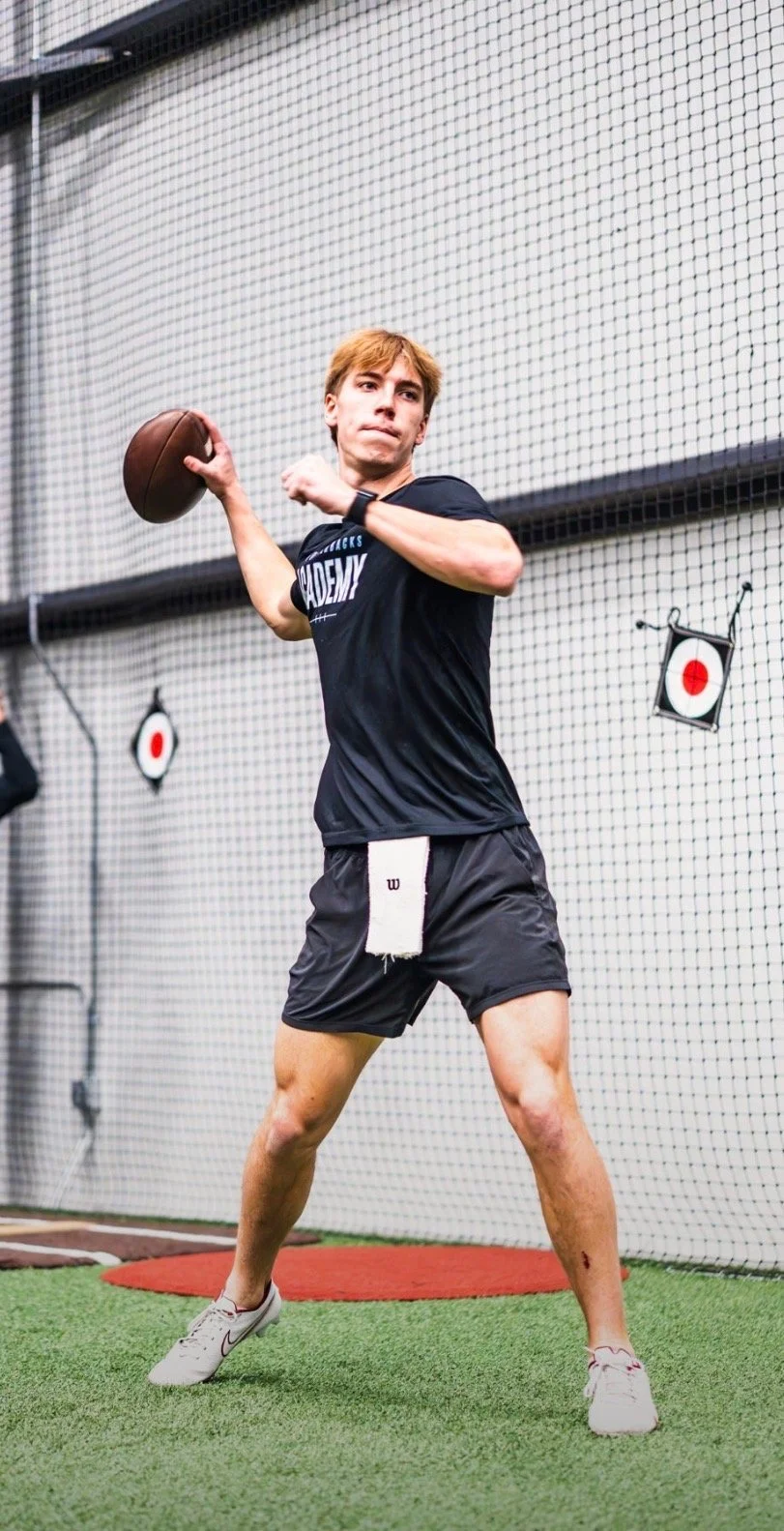 A young man in athletic clothes prepares to throw a football indoors, standing on artificial turf with a netted wall and target signs in the background.