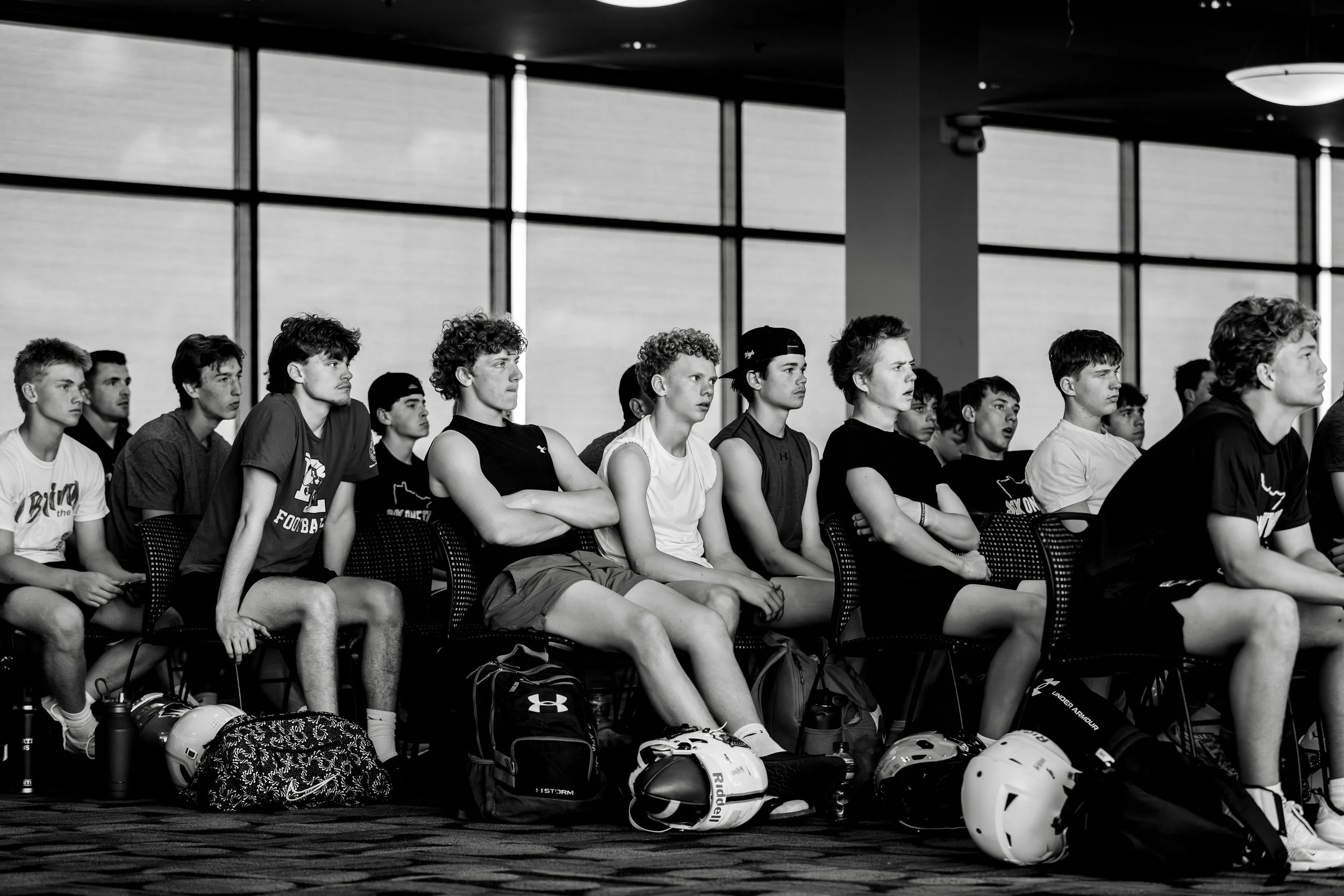 A group of young athletes sitting on chairs in an indoor space with large windows, some wearing sports gear and helmets on the floor in front of them, attentively watching something.