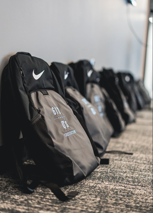 Row of black and gray Nike sports backpacks with personalized initials and names, placed on a carpeted floor.