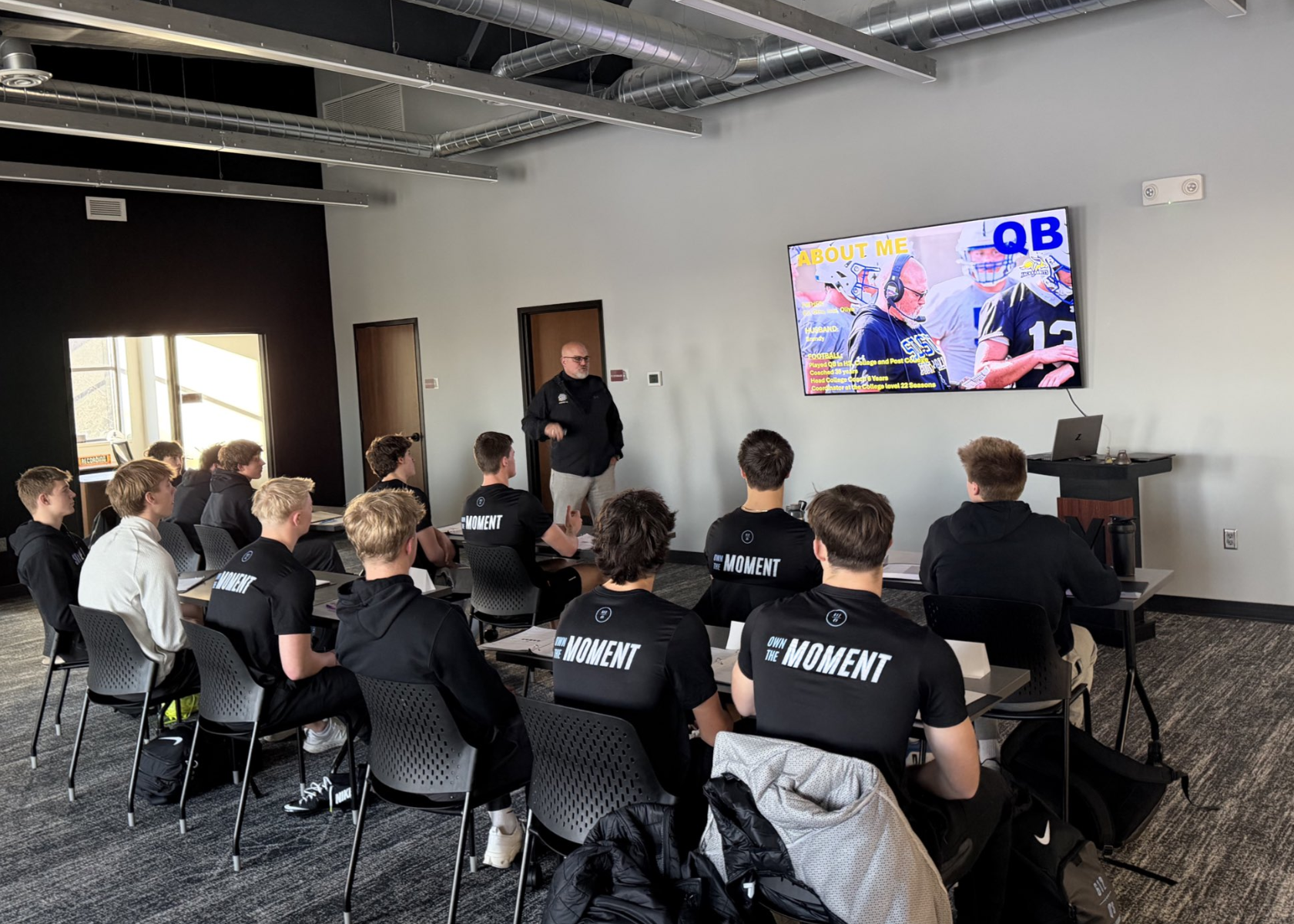 A classroom filled with young men attentively listening to a man giving a presentation. The students are wearing black shirts with the text 'OWN THE MOMENT' on the back. There is a large screen displaying a slide about a football player, with text including 'ABOUT ME' and 'QB'. The room has a modern design with exposed ductwork on the ceiling and large windows letting in natural light.