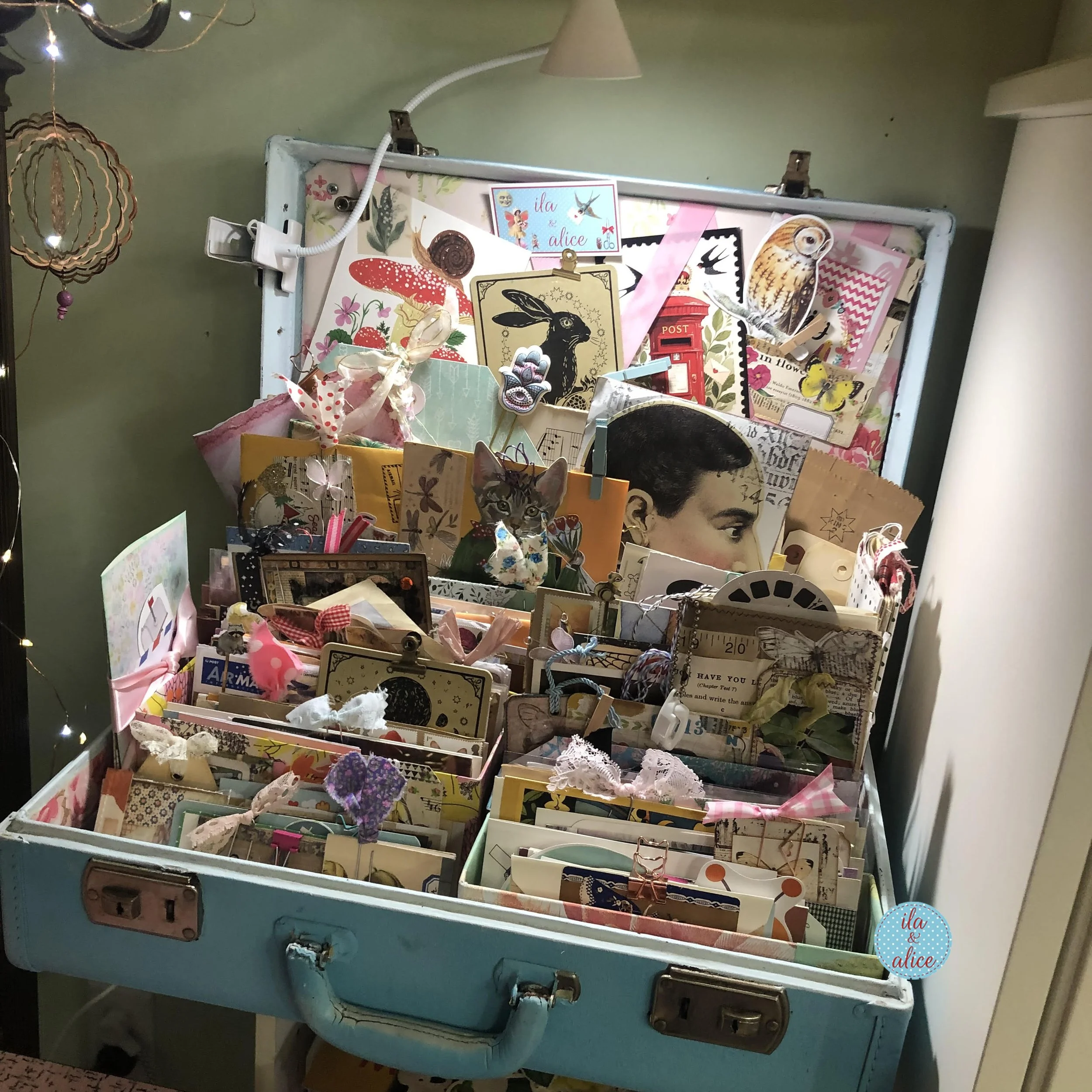 A vintage blue suitcase displayed in a craft room corner and filled with sorted happy mail, postcards, envelopes, tags, and paper keepsakes.