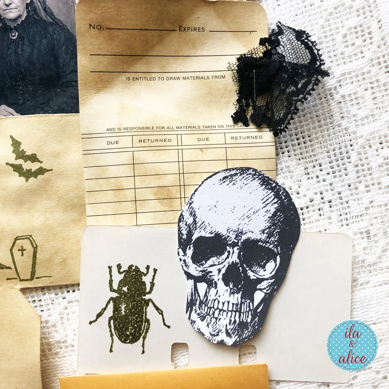 Gothic paper ephemera featuring a black-and-white skull cutout, stamped beetle card, coffee-dyed library card, bats, a coffin and a small piece of black lace.