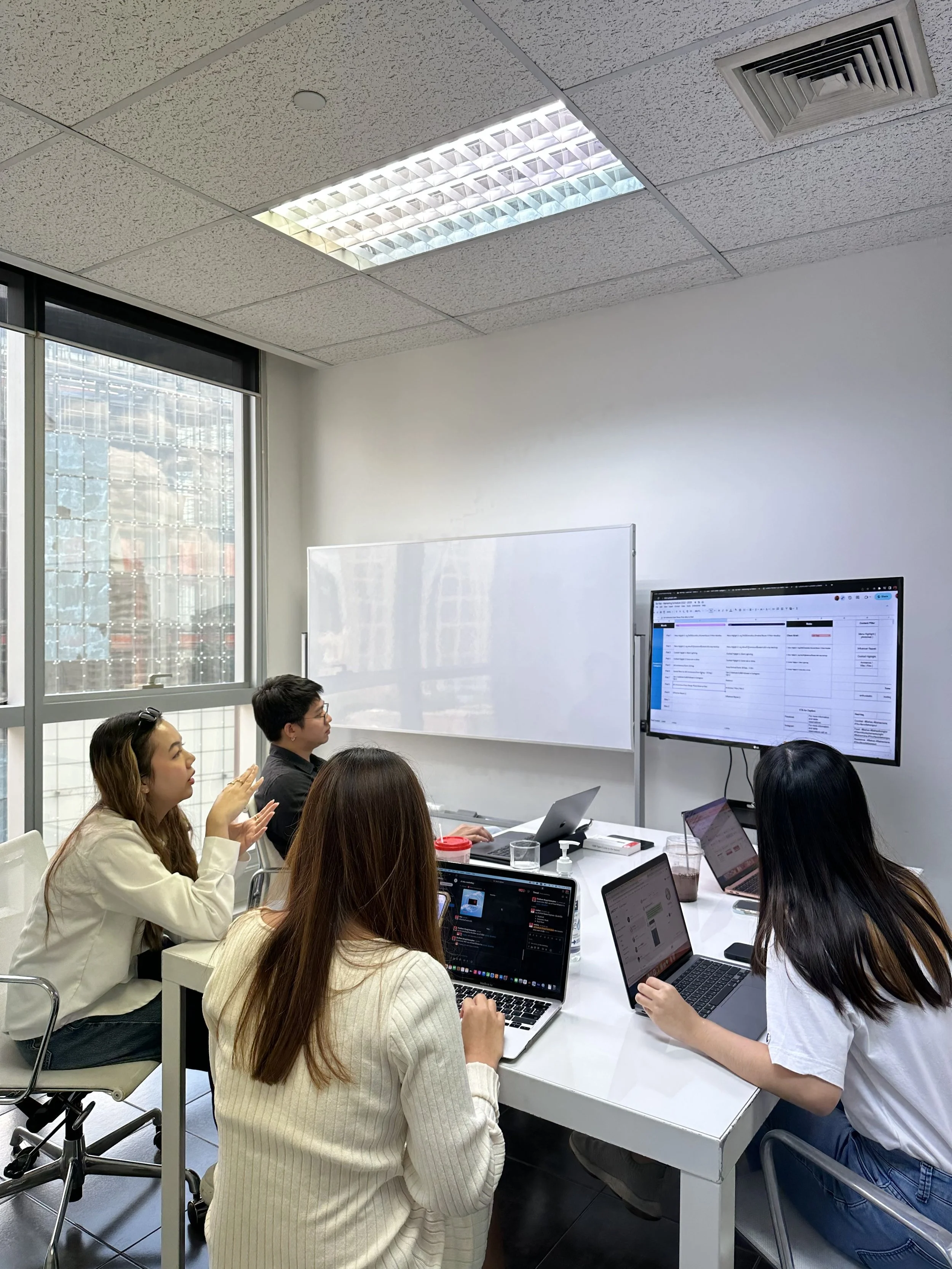 A group of four people in a meeting room with laptops, a large monitor displaying a spreadsheet, and a whiteboard. They are engaged in a discussion.