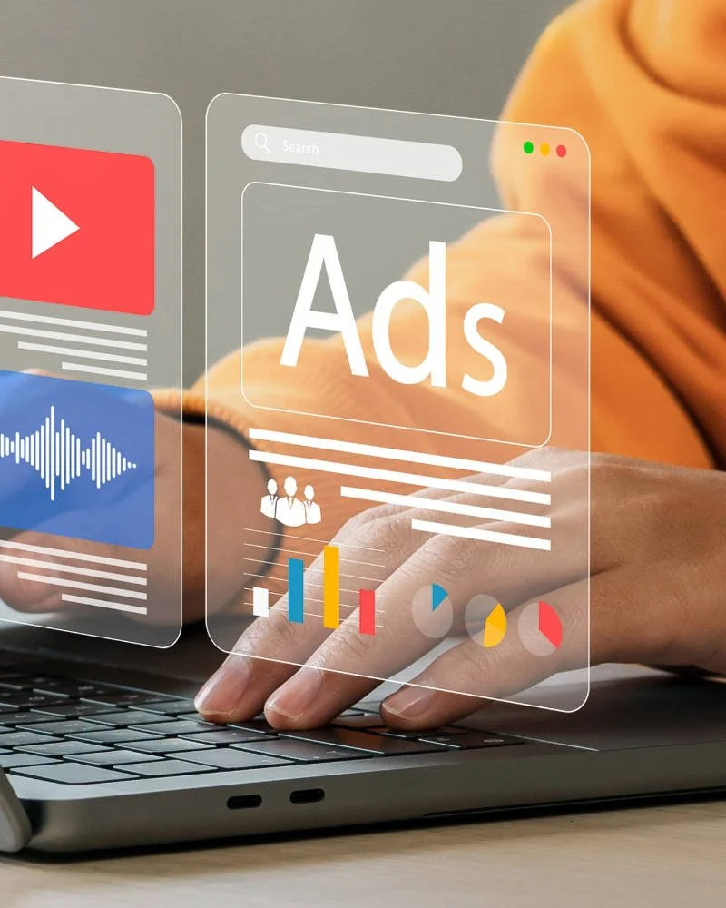 A person typing on a laptop with digital overlay graphics, including a large 'Ads' label, search bar, video play button, sound wave, charts, and user icons, representing online advertising and digital marketing.