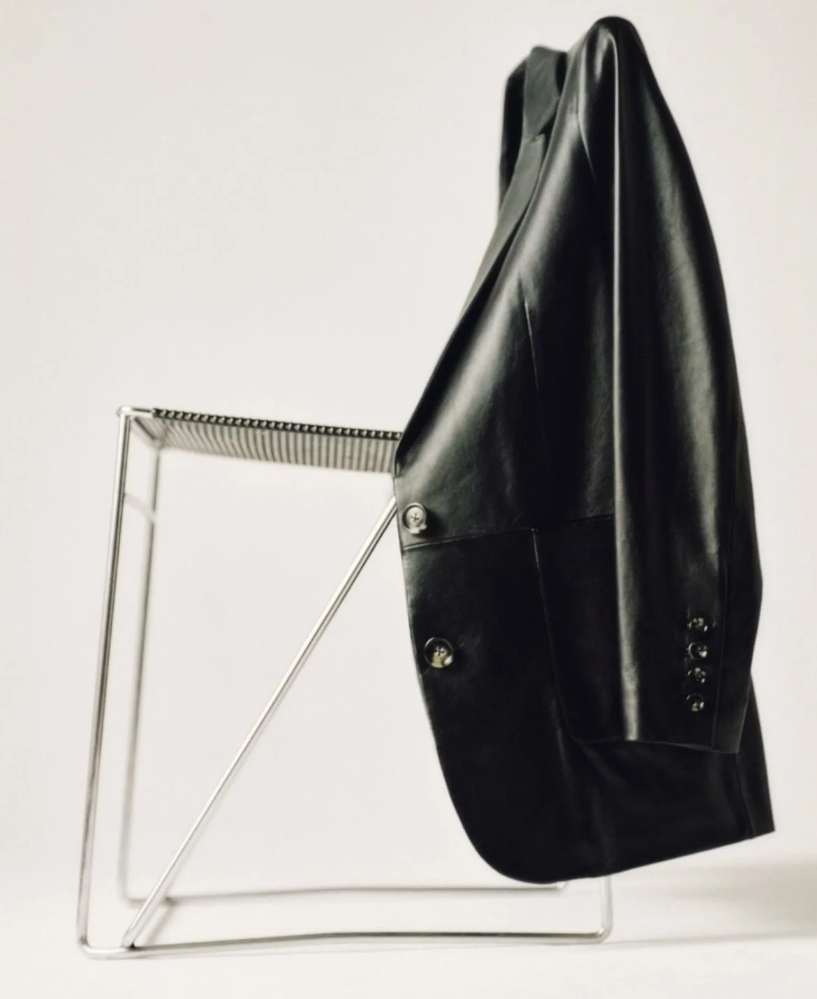 A sculptural black leather blazer casually hung over a modern metal chair — evoking bold silhouettes and wardrobe edge for a sleek, elevated look.