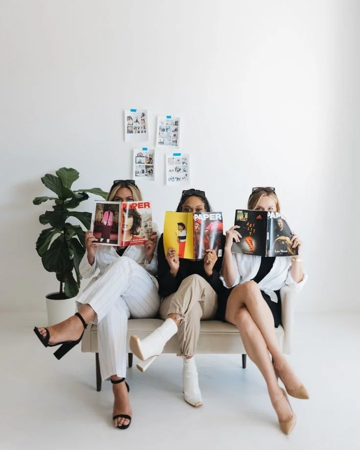 A stylish trio of women dressed in chic business-casual outfits, sitting on a modern beige couch while holding up fashion magazines. Their playful yet polished demeanor, combined with a minimal studio setting, reflects a contemporary, trend-conscious