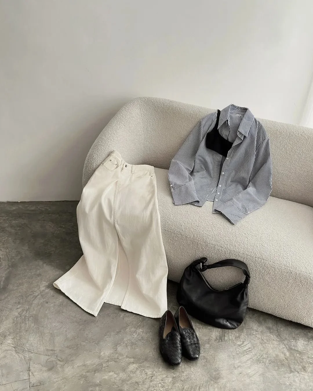 A soft-toned outfit styled on a boucle sofa featuring a striped shirt, wide-leg ivory trousers, woven black flats, and a black leather bag — representing everyday style with a refined, minimal aesthetic.