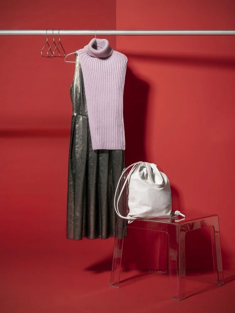 A shimmering metallic dress paired with a lilac knit turtleneck and a white bucket bag, styled against a bold red backdrop — showcasing statement styling for personality-driven wardrobes.