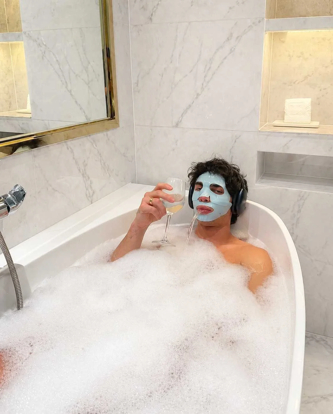 Lifestyle image of a man relaxing in a bath with a sheet mask, representing modern men’s grooming, skincare routines, and self-care rituals