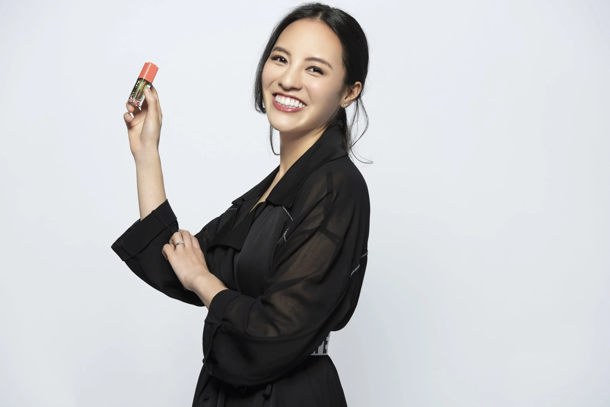 A woman with black hair, smiling and holding a small container with a coral-colored lid, standing against a plain white background.