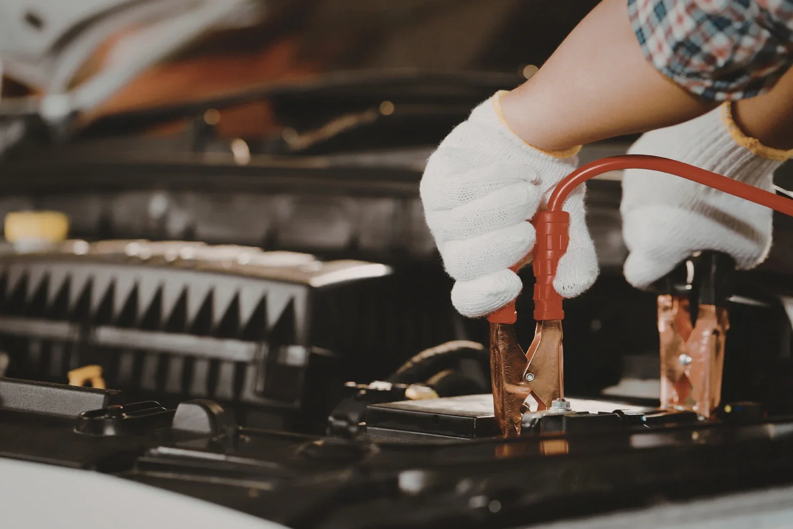 Person wearing protective gloves connecting jumper cables to a car battery under an open hood for a battery jumpstart.