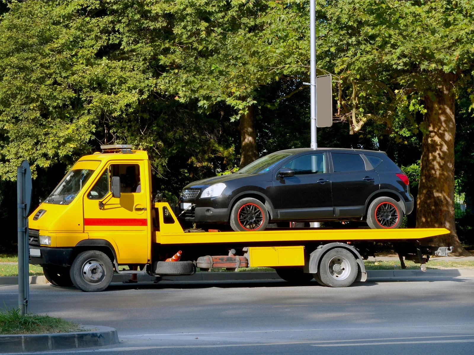 Yellow flatbed tow truck transporting a black SUV on a roadside lined with large green trees during daylight.