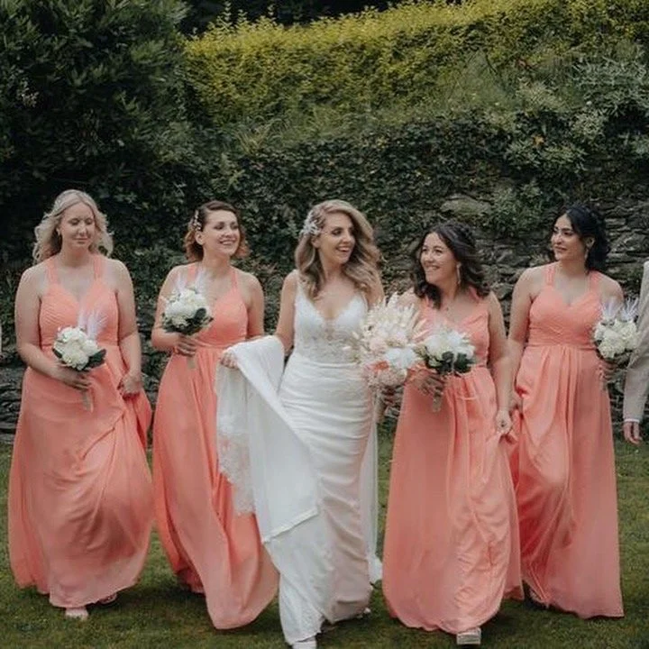 A bride in a white wedding dress walking outdoors with five bridesmaids in peach dresses, each holding a bouquet of flowers.