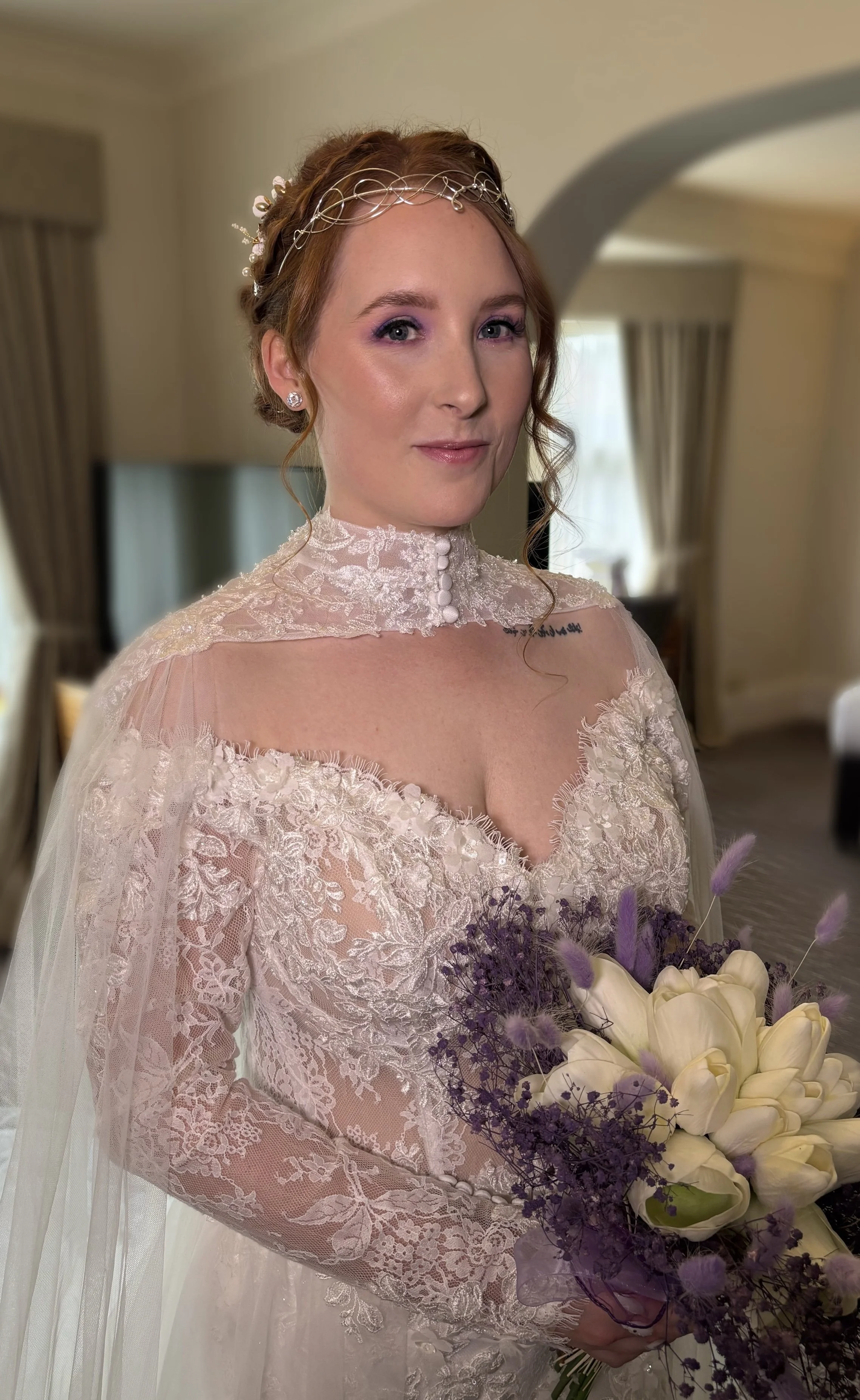 A woman in a wedding dress holding a bouquet of white calla lilies and purple flowers, standing in a room with curtains and soft lighting.