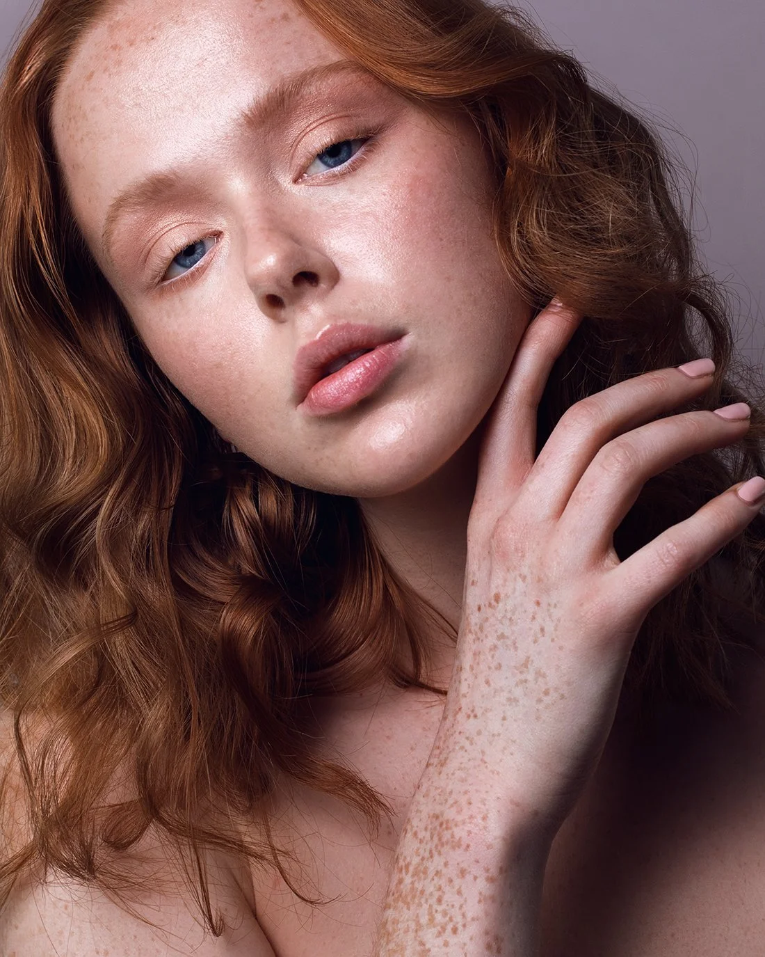 Close-up of a young woman with red hair and freckles, touching her neck with her hand, with a neutral background.