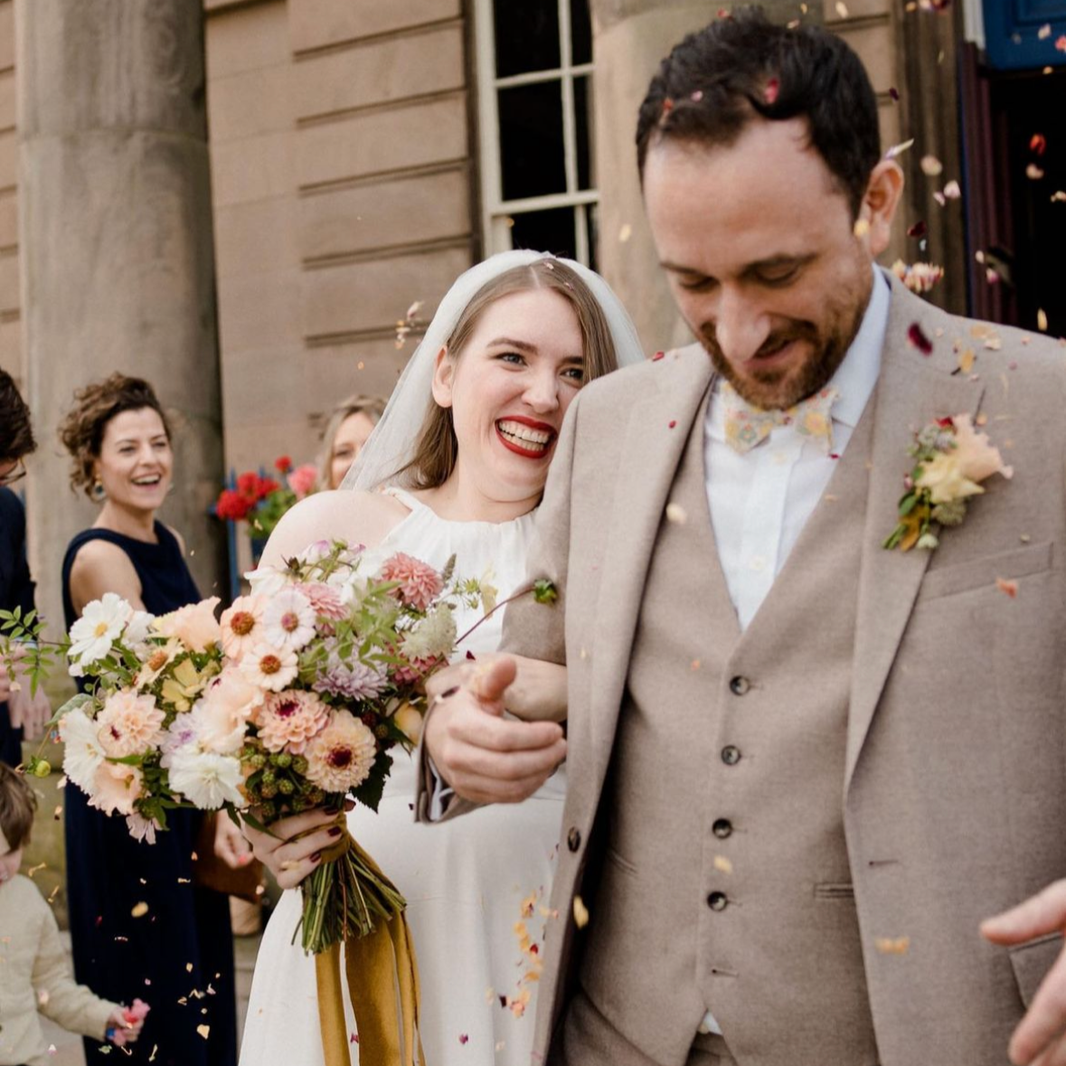 A bride and groom celebrating their wedding with friends and family outside, surrounded by flower petals and confetti.