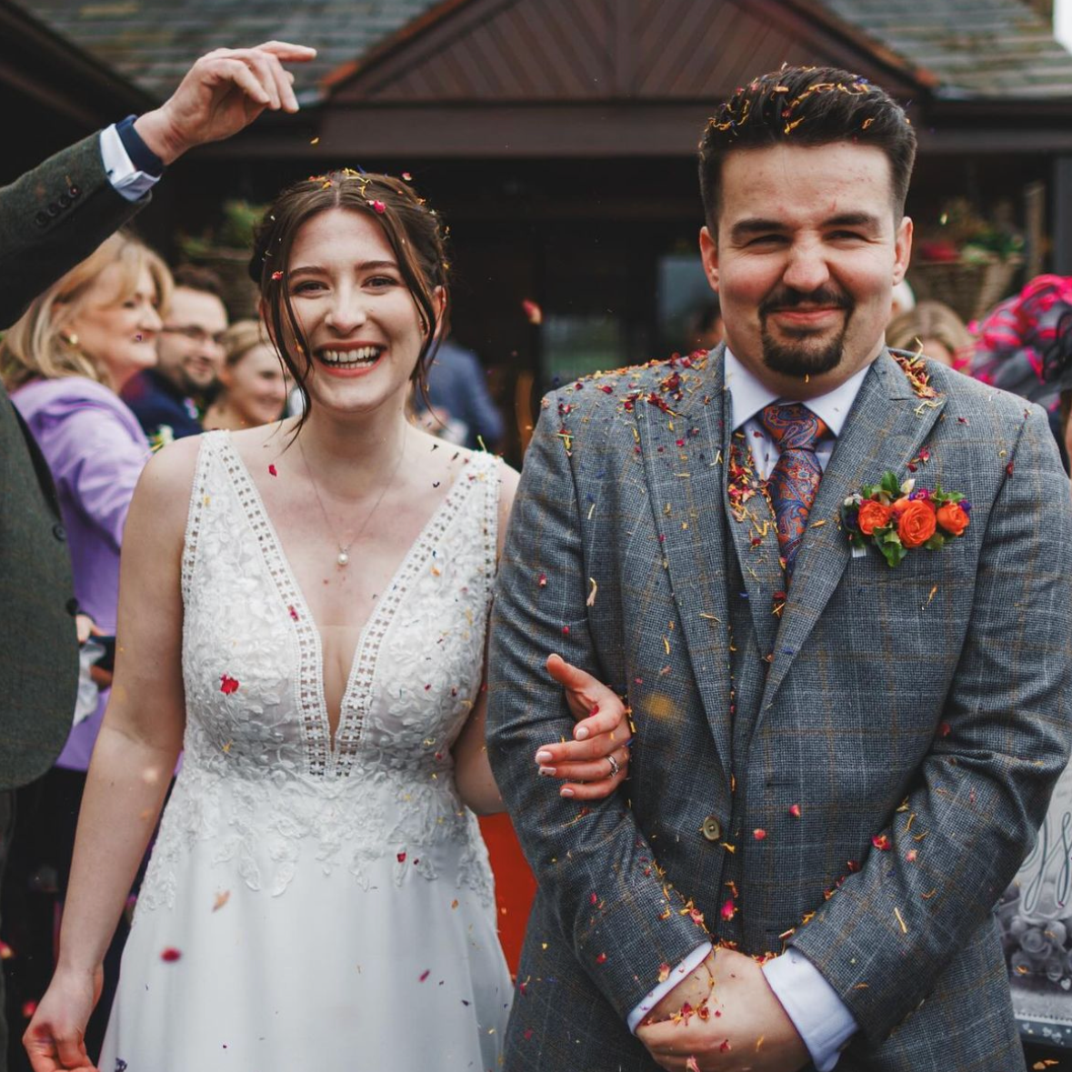 A photo of a smiling bride in a white wedding dress and a groom in a grey suit, standing outside a house during a celebration with confetti falling around them.