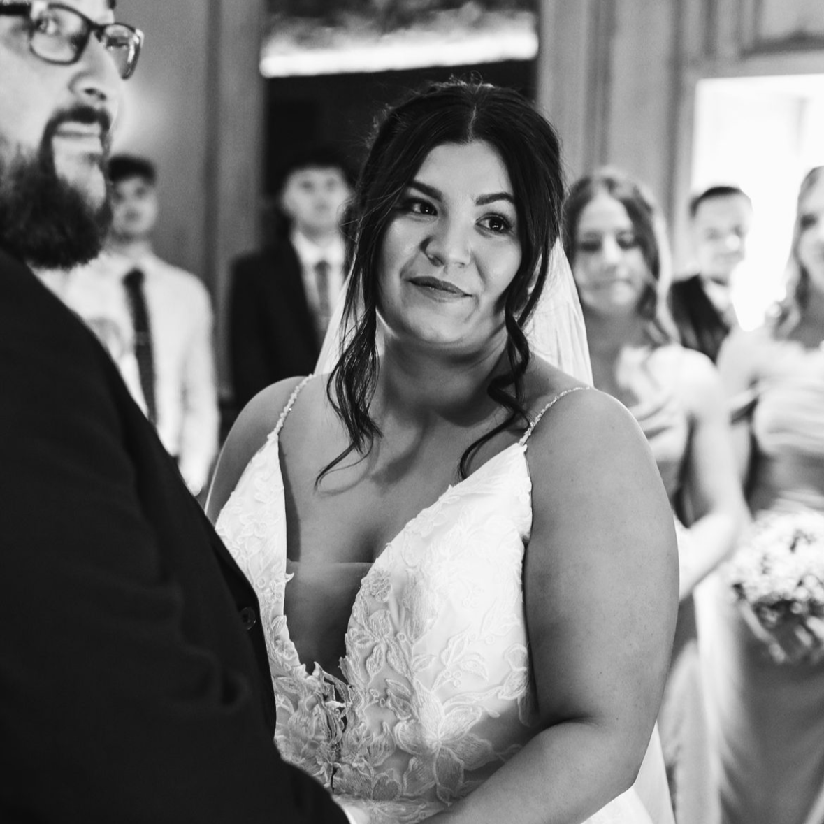 Black and white photo of a bride and groom holding hands during their wedding ceremony, with guests in the background, indoors.