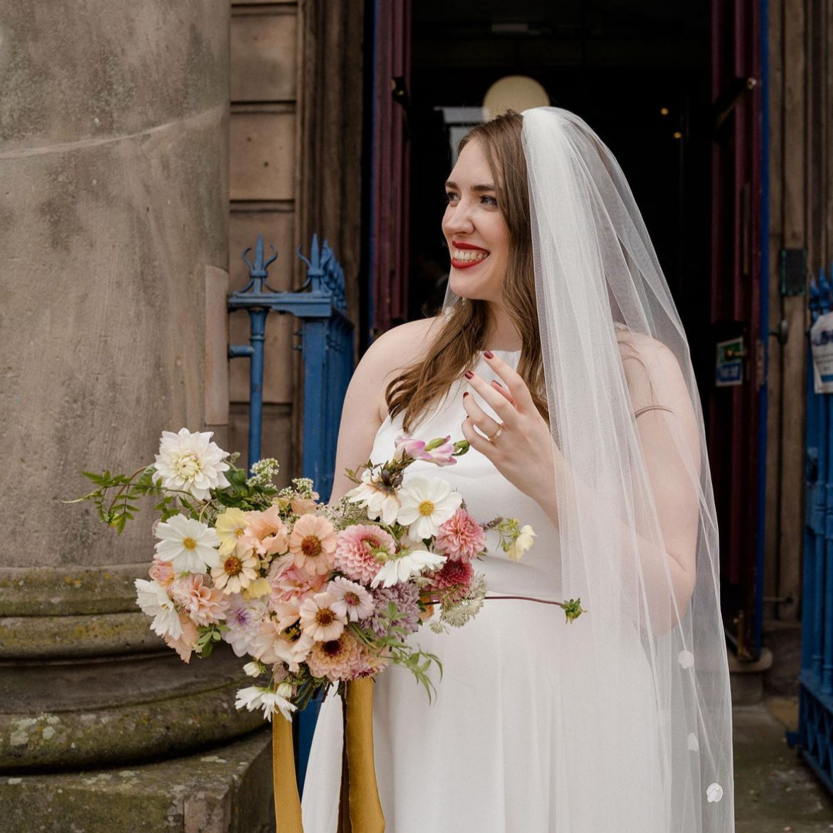 A woman in a wedding dress holding a bouquet of flowers, standing outdoors near a stone column and a blue gate. She is smiling and wearing a veil.