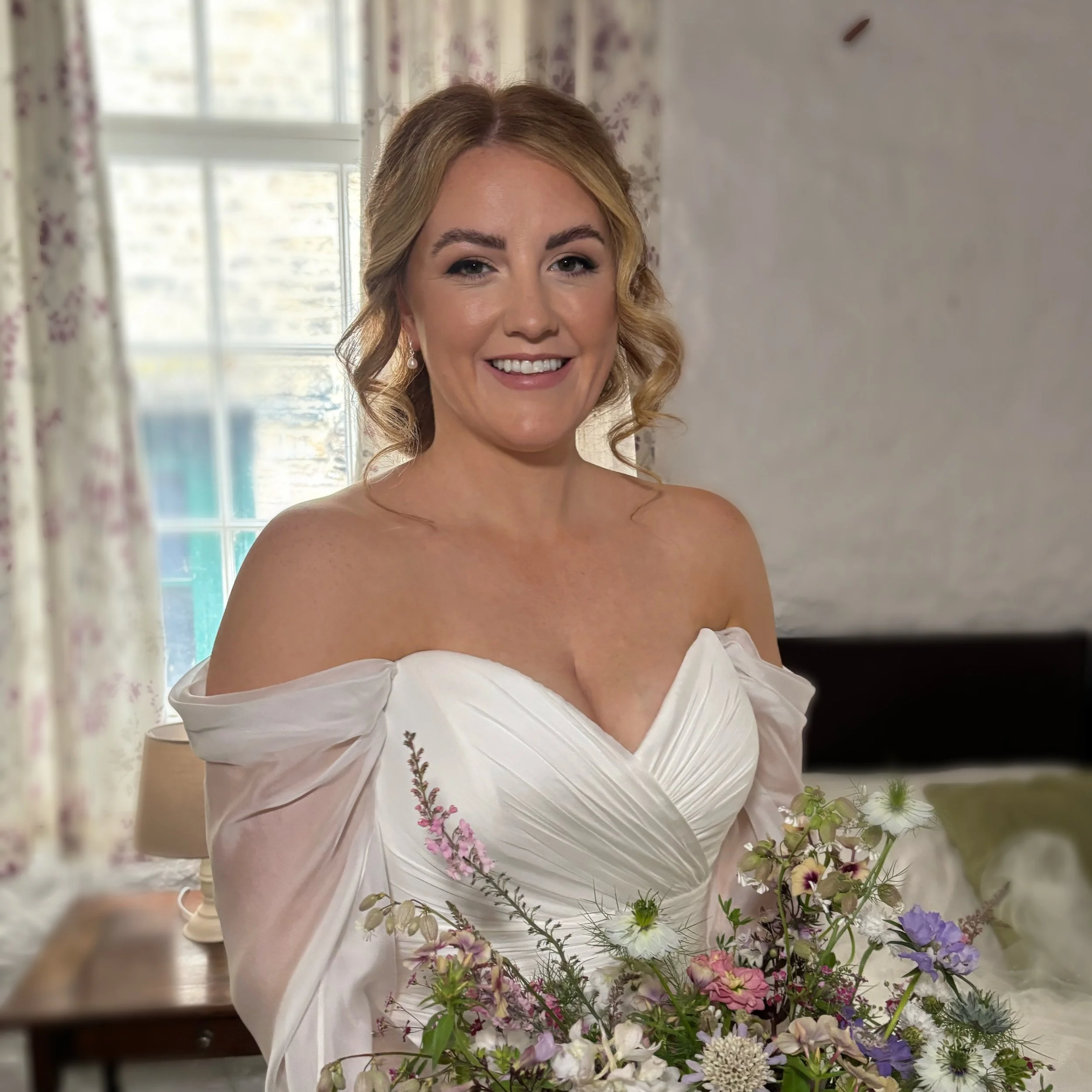 A woman in a white off-the-shoulder wedding dress holding a bouquet of flowers, smiling near a window with floral curtains.