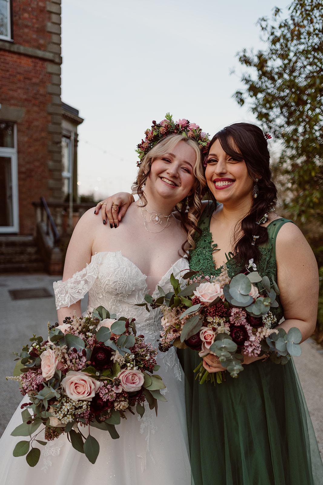 Two women in wedding attire standing outdoors, smiling, holding bouquets of flowers, one with a floral crown, in front of a brick building and trees.