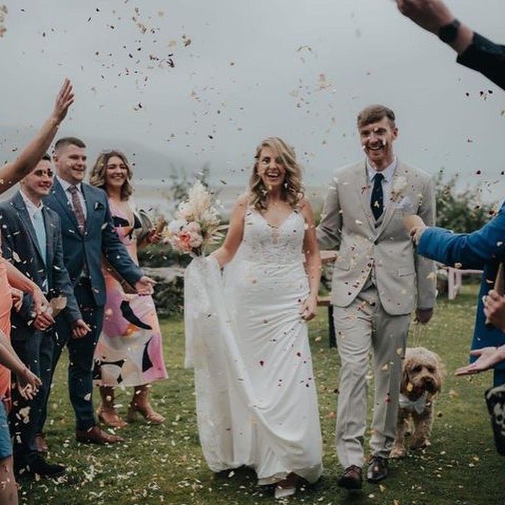 Bride and groom walking outdoors after wedding ceremony, surrounded by friends throwing confetti, with a dog nearby, on a cloudy day.