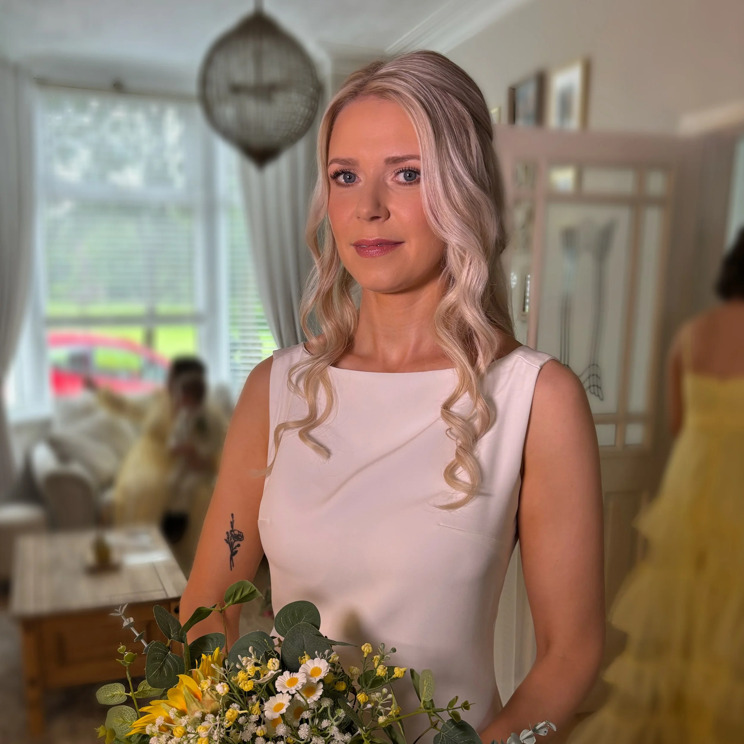 A woman with wavy blonde hair wearing a white dress holds a bouquet of yellow and white flowers in a room decorated for a wedding or special occasion.