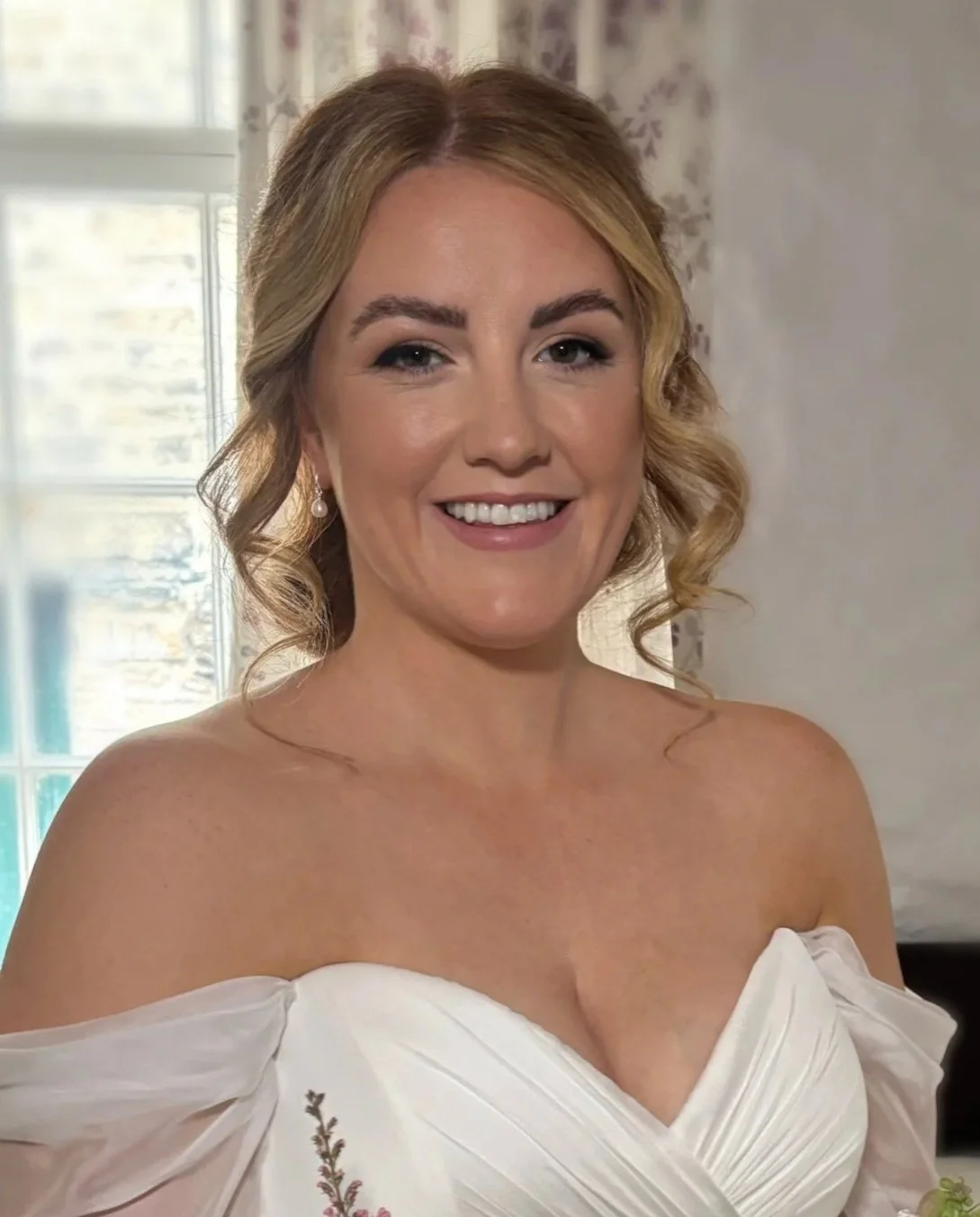 Smiling woman with blonde curly hair wearing pearl earrings and a white off-shoulder dress, standing in front of a window with floral curtains.