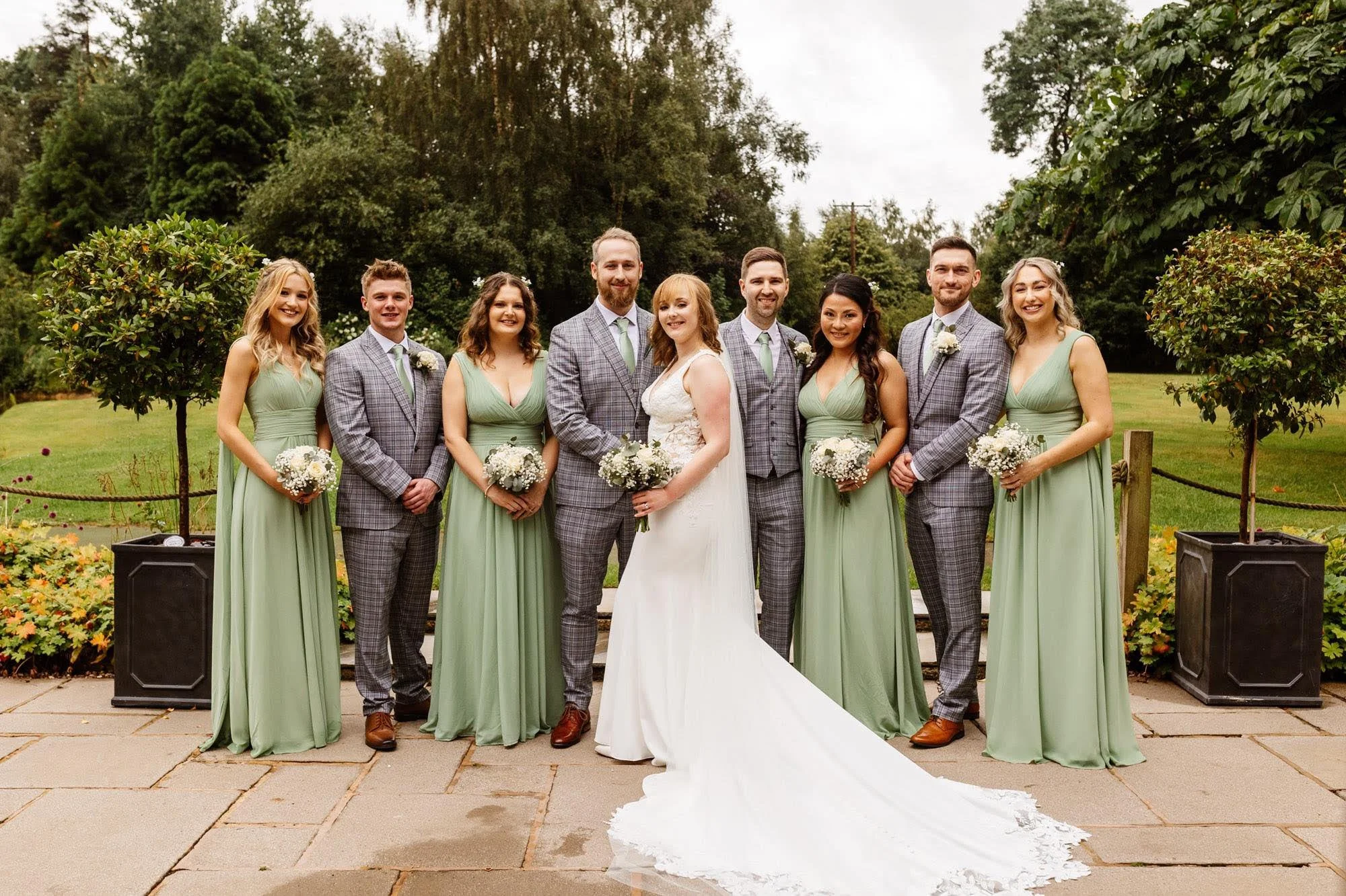 Wedding party poses outdoors on a brick patio, with the bride in a white dress and veil holding a bouquet, surrounded by bridesmaids in light green dresses and groomsmen in gray plaid suits, all smiling.