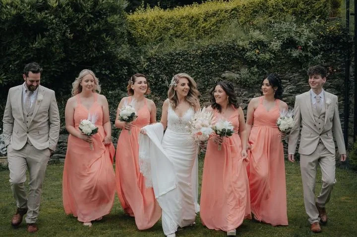 Bride and bridal party walking outdoors in front of greenery, including five women in peach dresses holding bouquets and two men in beige suits.