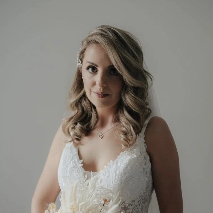 A bride with wavy blonde hair, wearing a white lace wedding dress and a heart-shaped necklace, holding a bouquet of flowers.