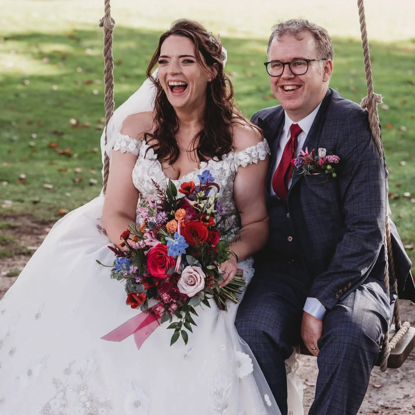 A bride and groom sitting on a swingset, smiling and laughing outdoors. The bride is holding a colorful bouquet of flowers and wearing a white wedding dress. The groom is dressed in a checkered suit with a red tie and glasses.