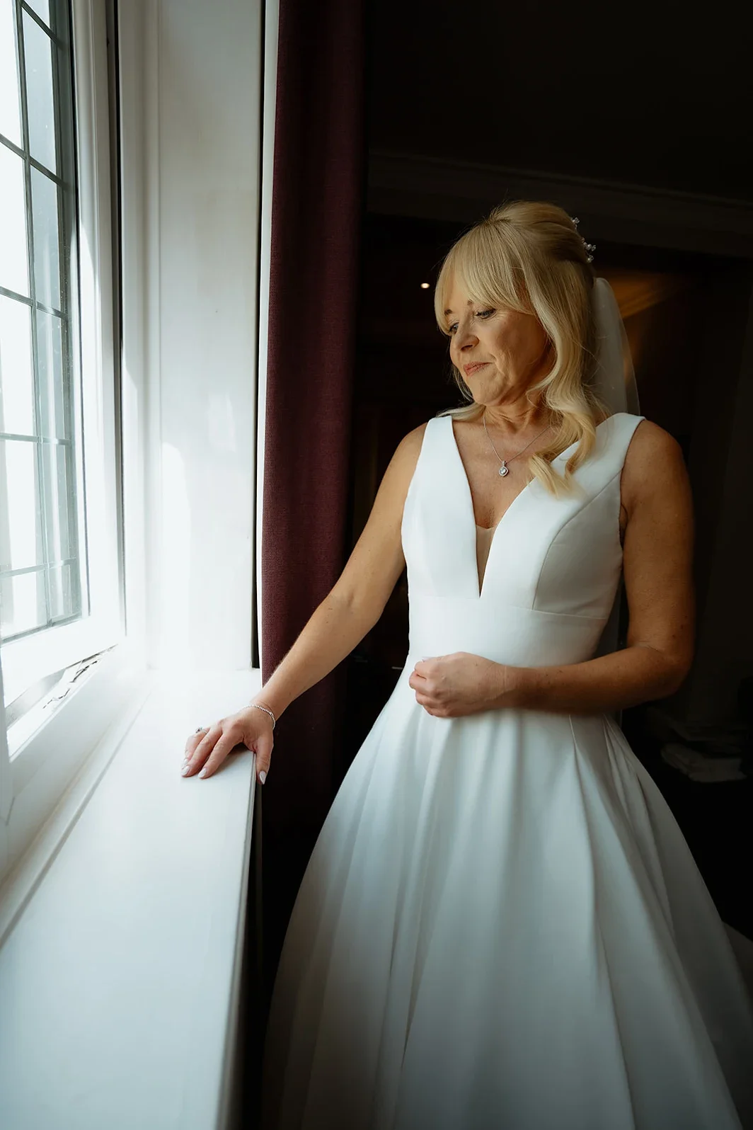A bride in a white wedding dress, standing by a window, looking outside with a gentle smile and a contemplative expression.