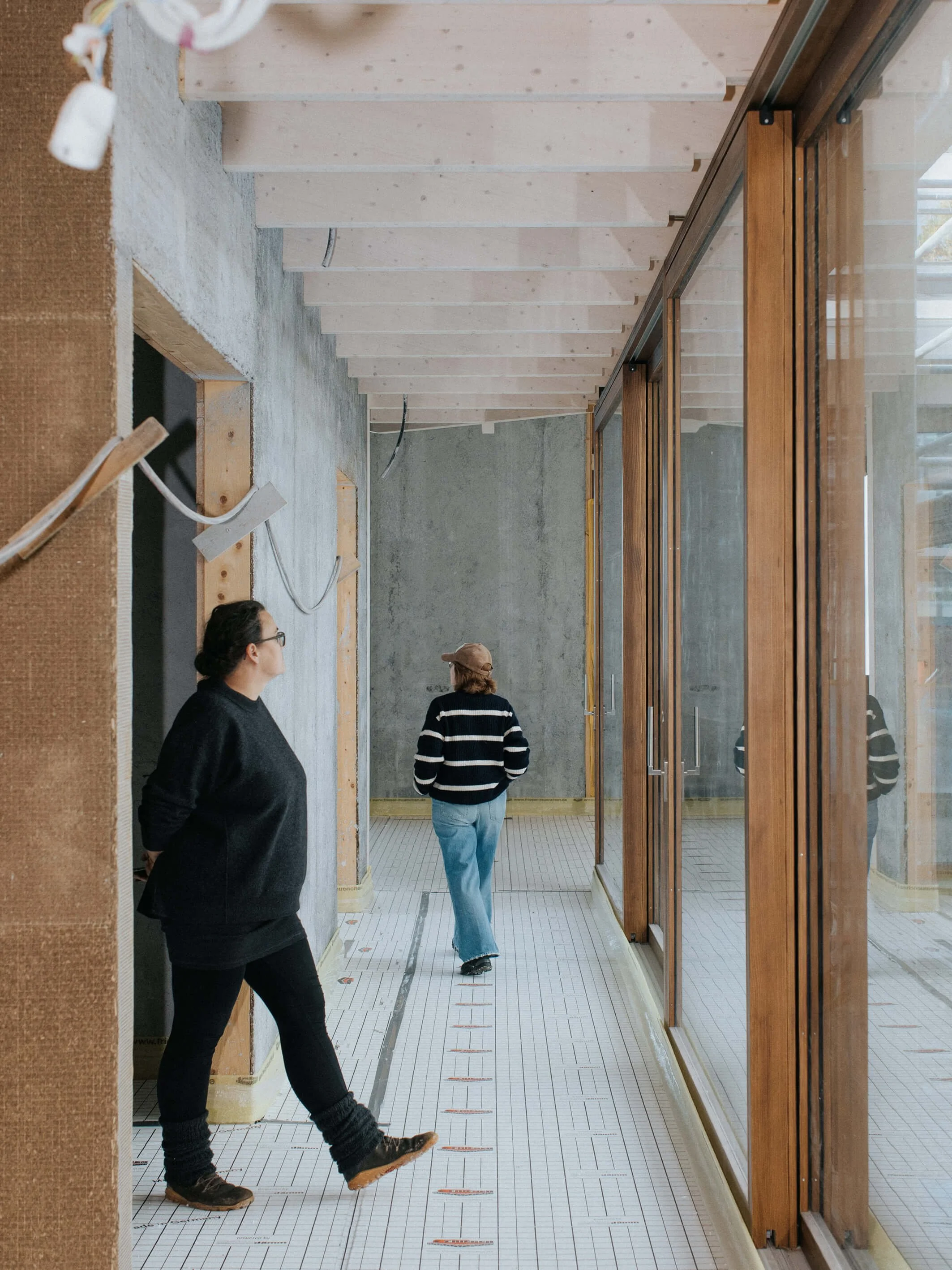 Innenansicht eines Bau- oder Renovierungsprojekts mit zwei Frauen, die den Raum besichtigen. Der Raum hat ein rohes, unvollendetes Erscheinungsbild mit sichtbarem Holz und Beton.