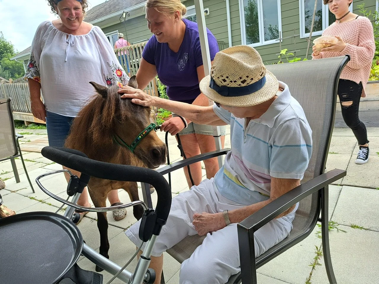 Older man wearing a straw hat, sitting in a patio chair and petting a small horse, with three young women nearby smiling and holding snacks, in a backyard with a wooden deck.