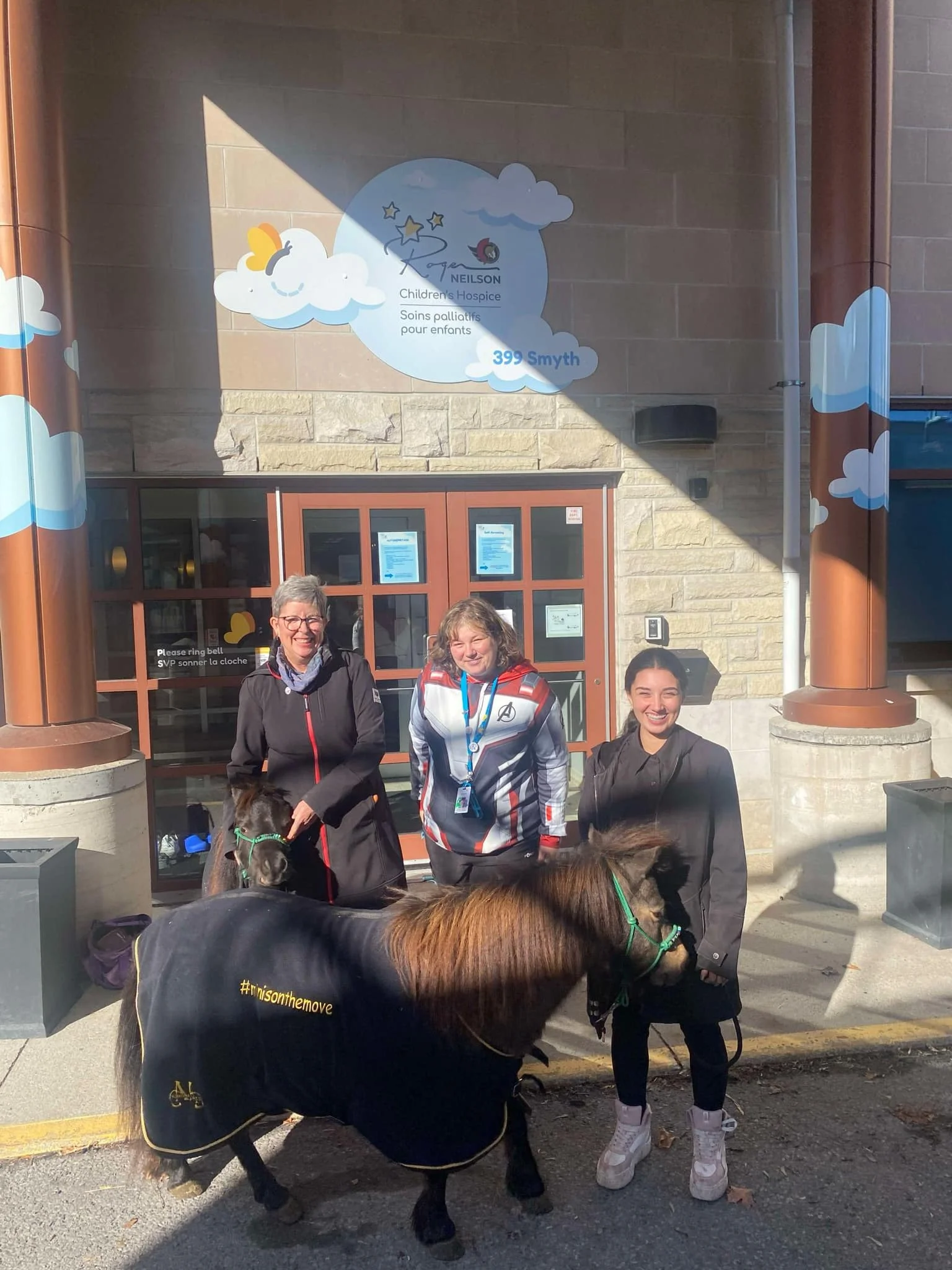 Three women standing outside a building with two small ponies, one black and one brown, wearing blankets. The building has a sign for a children's hospice and decorative clouds and stars. The women are smiling, and the woman on the right is holding the brown pony's leash.