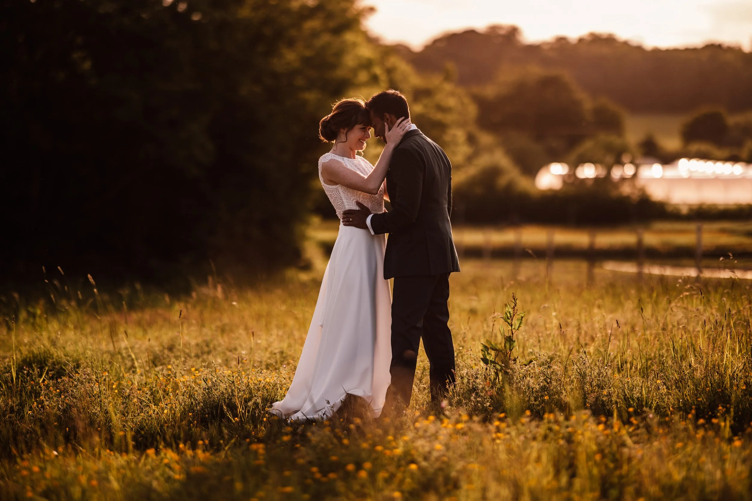 A bride and groom embracing each other in a field at sunset, with trees and a distant building in the background.