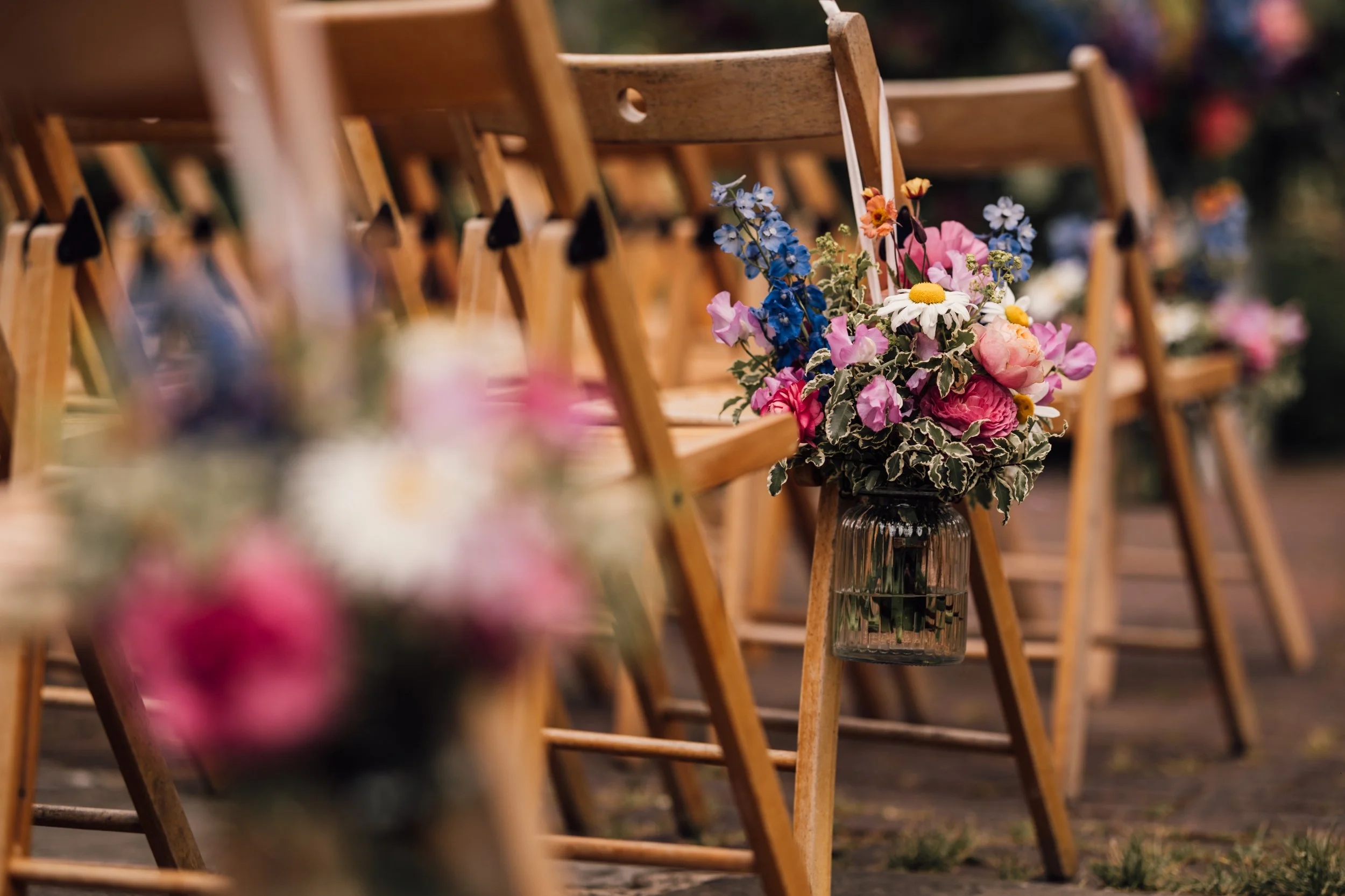 Wooden chairs arranged in rows with floral arrangements hanging from the sides, including pink, white, and blue flowers and greenery, at an outdoor event.
