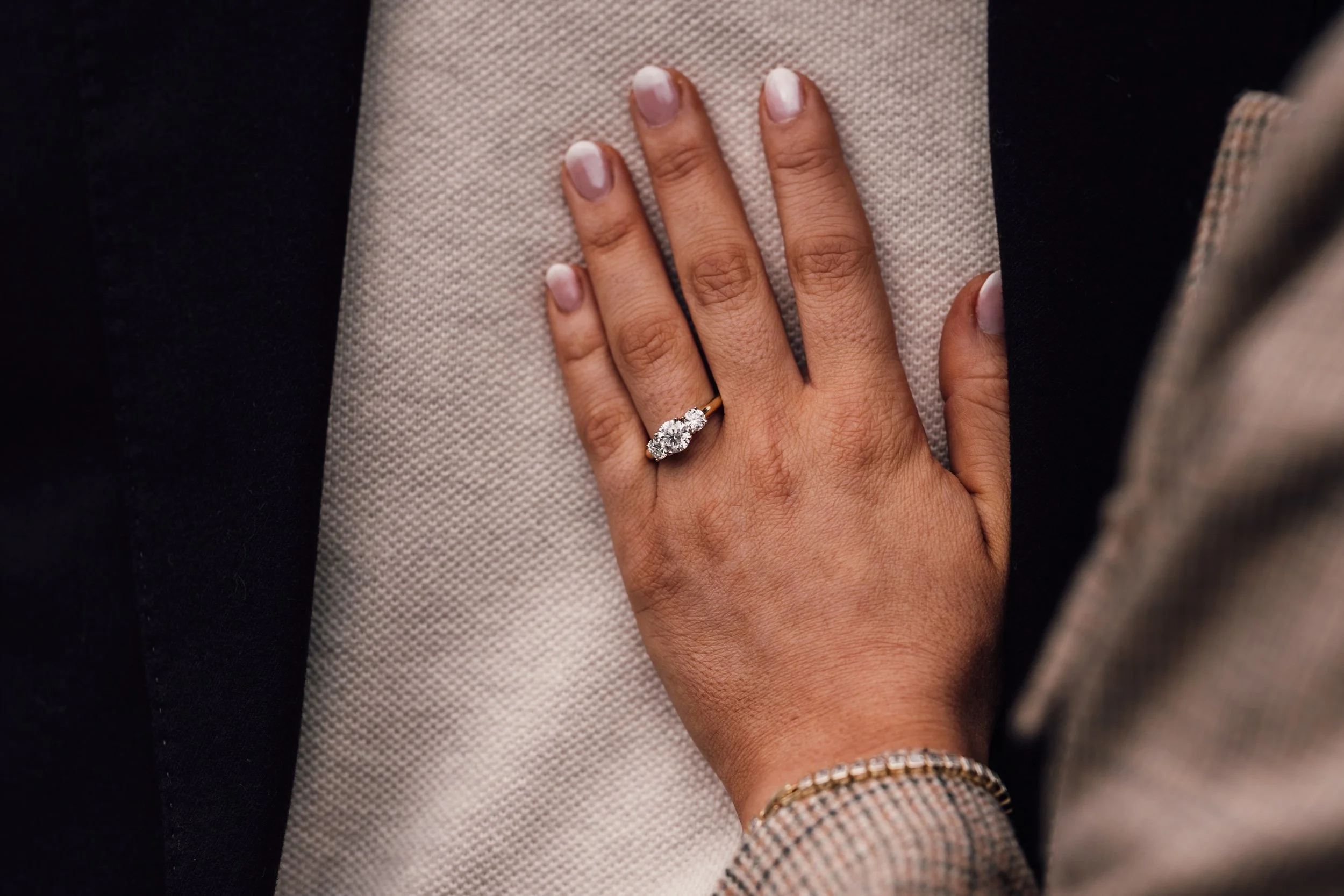 A woman's hand with a diamond engagement ring resting on a surface, wearing a bracelet and a plaid jacket.