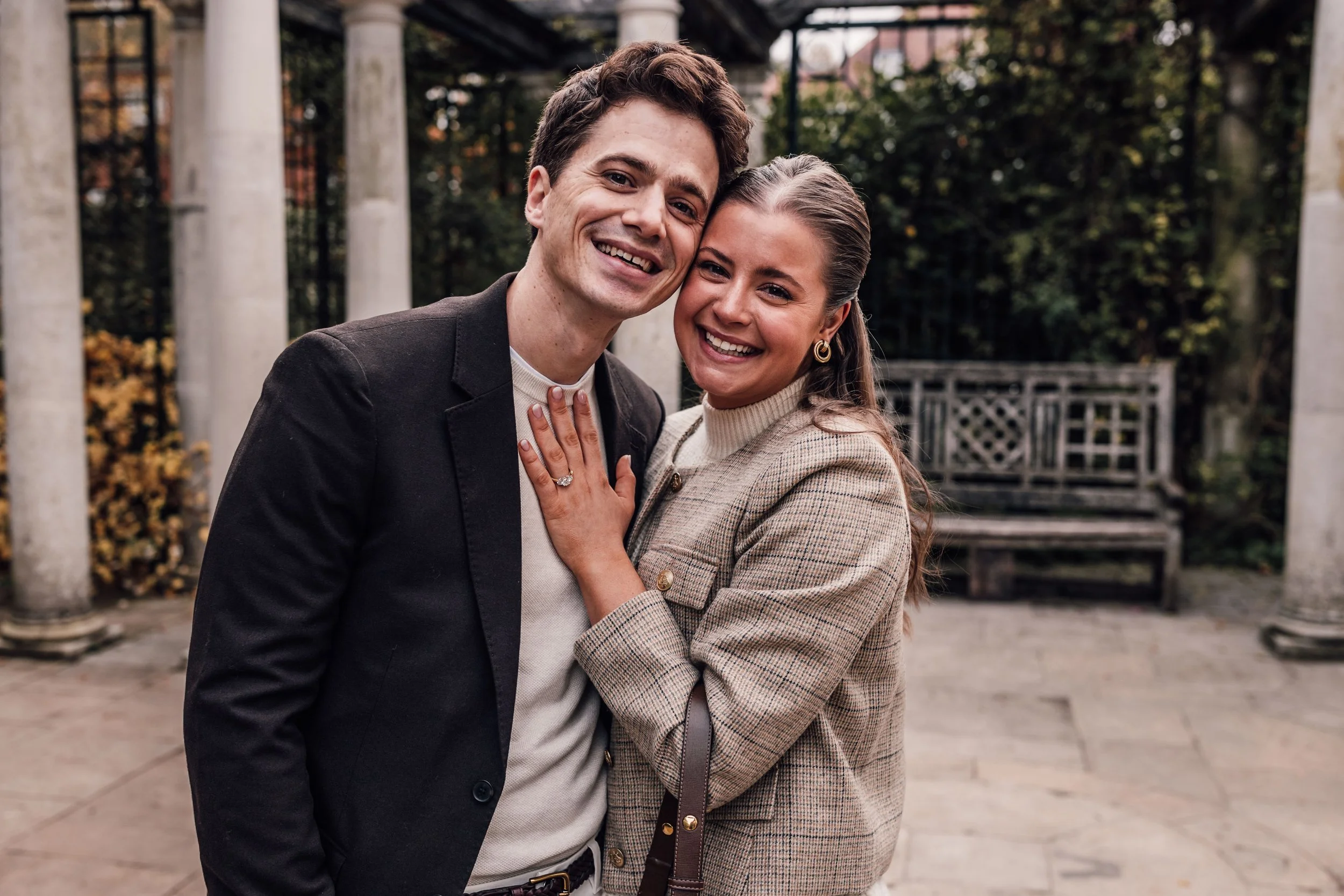 A smiling couple, a man and a woman, standing close together outdoors, with the woman showing an engagement ring on her left hand. They are surrounded by trees, columns, and a bench in the background, suggesting a park or garden setting.