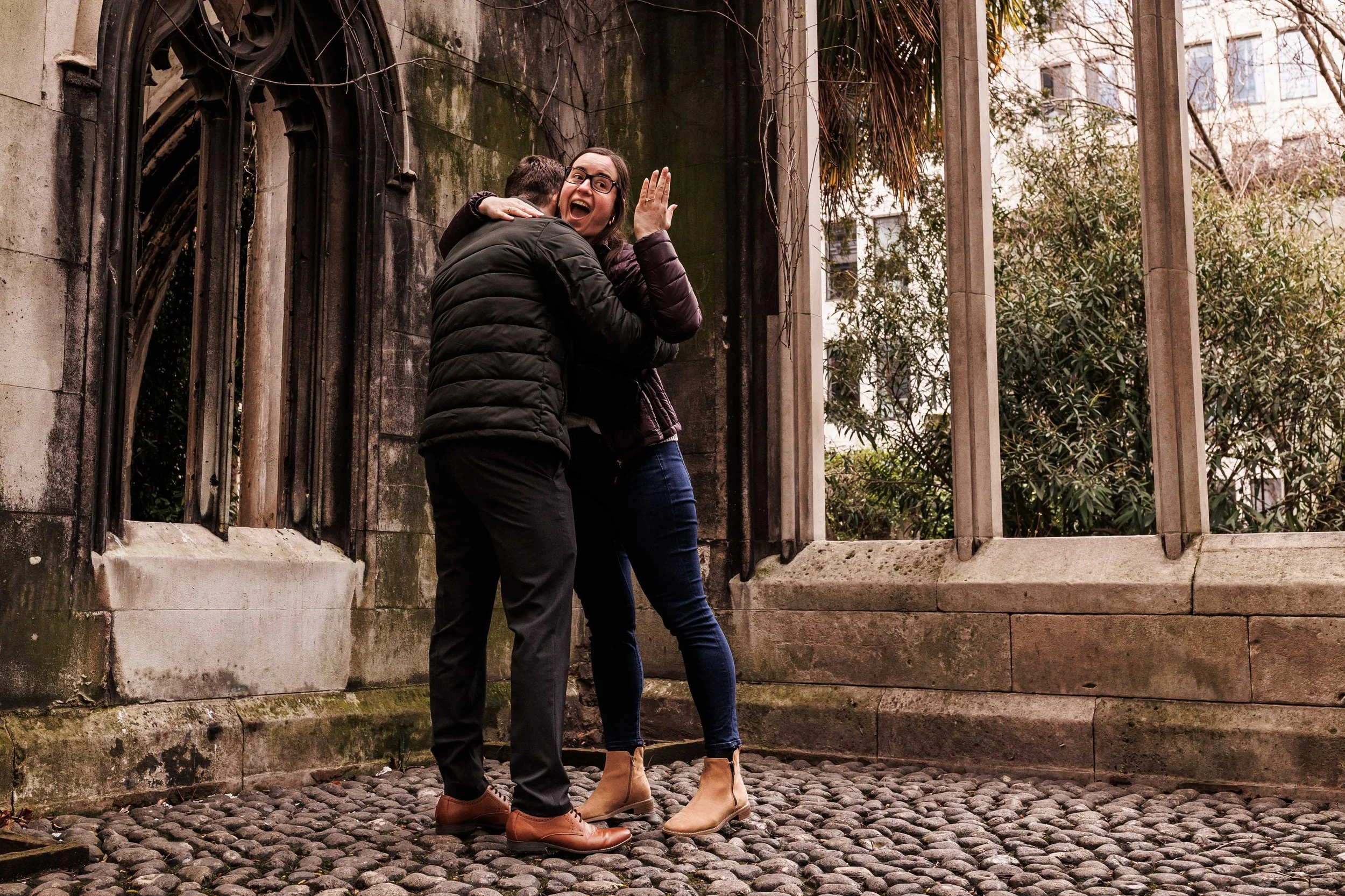 Two people hugging outdoors on cobblestone ground, woman with glasses and dressed in dark clothing smiling and waving, man with dark jacket hugging her, surrounding greenery and stone architecture in the background.