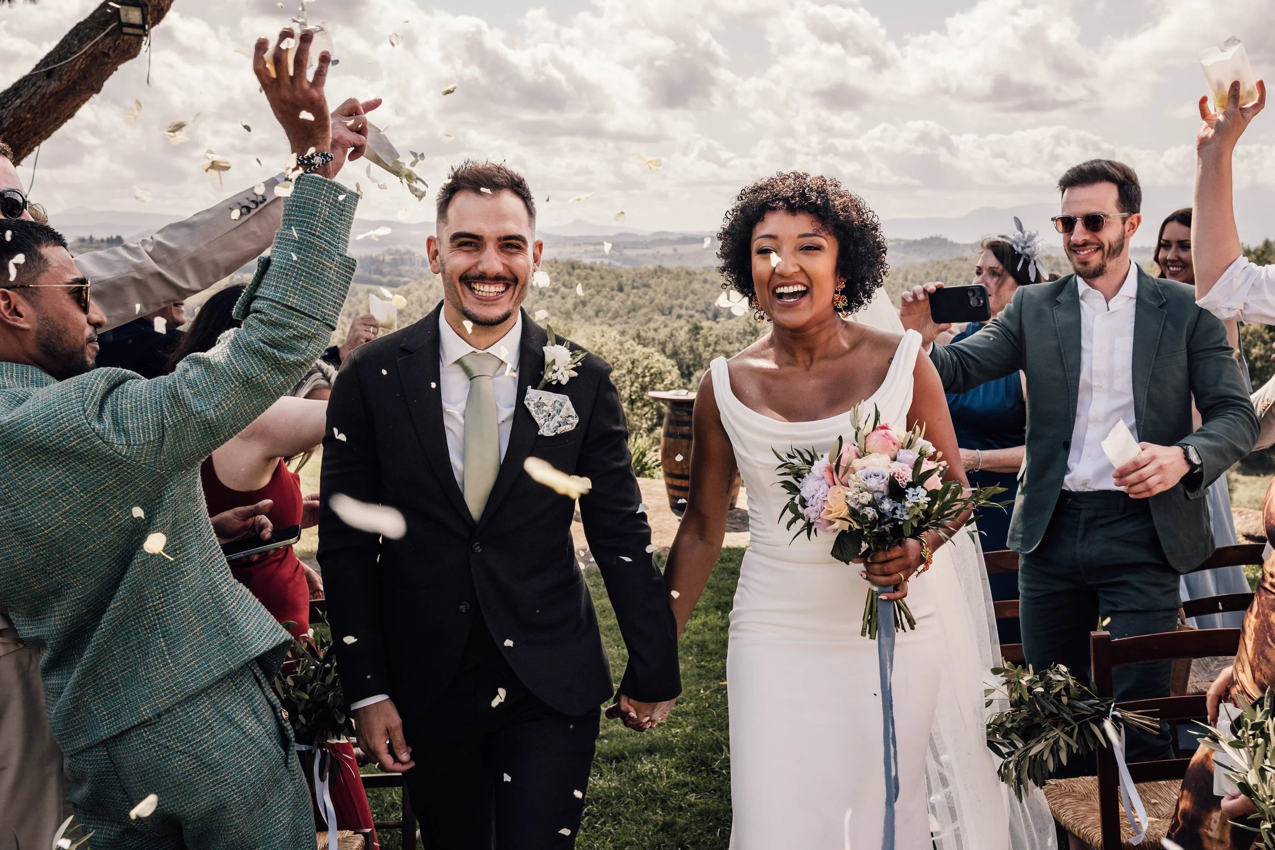 A joyful wedding celebration outdoors with a bride and groom walking hand in hand, surrounded by friends throwing confetti, with scenic mountains and cloudy sky in the background.