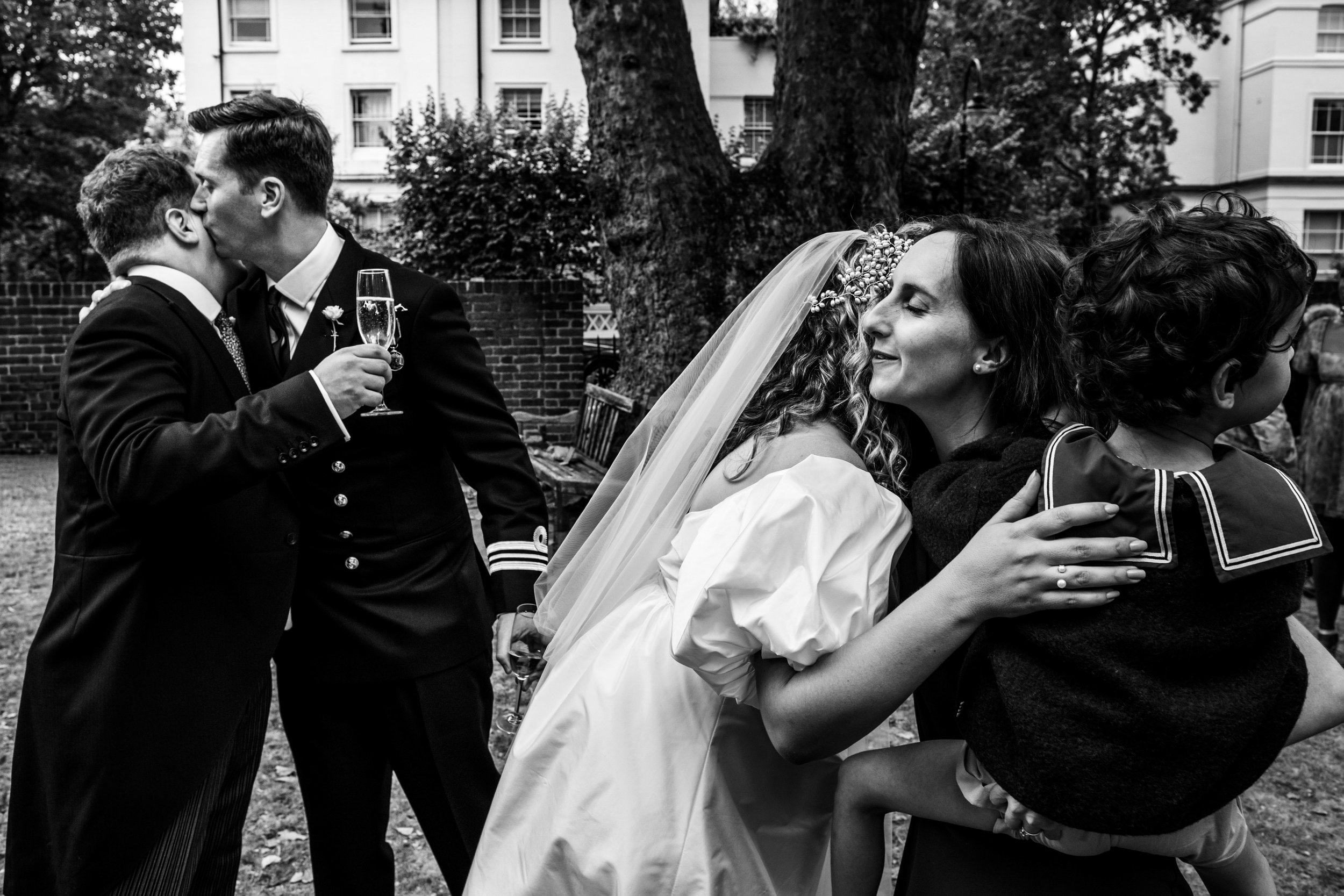 Black and white photo of a wedding scene with two couples hugging and kissing outdoors, with trees and buildings in the background.