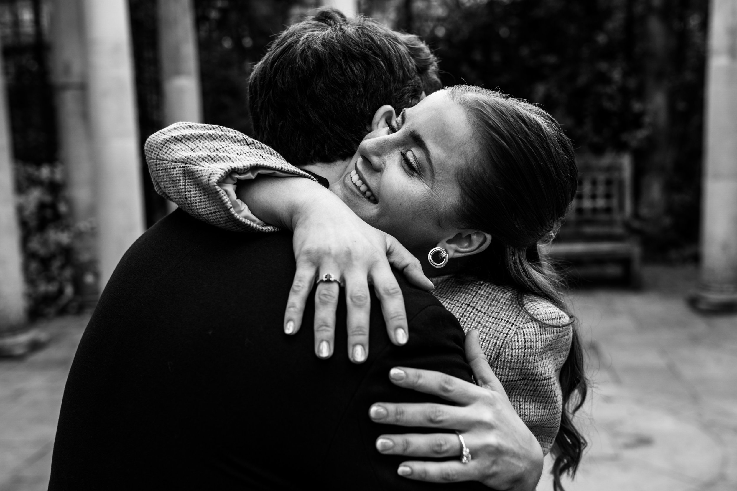A woman with long hair and earrings is hugging a man, smiling with her eyes closed. They are outdoors near trees, and the photo is in black and white.