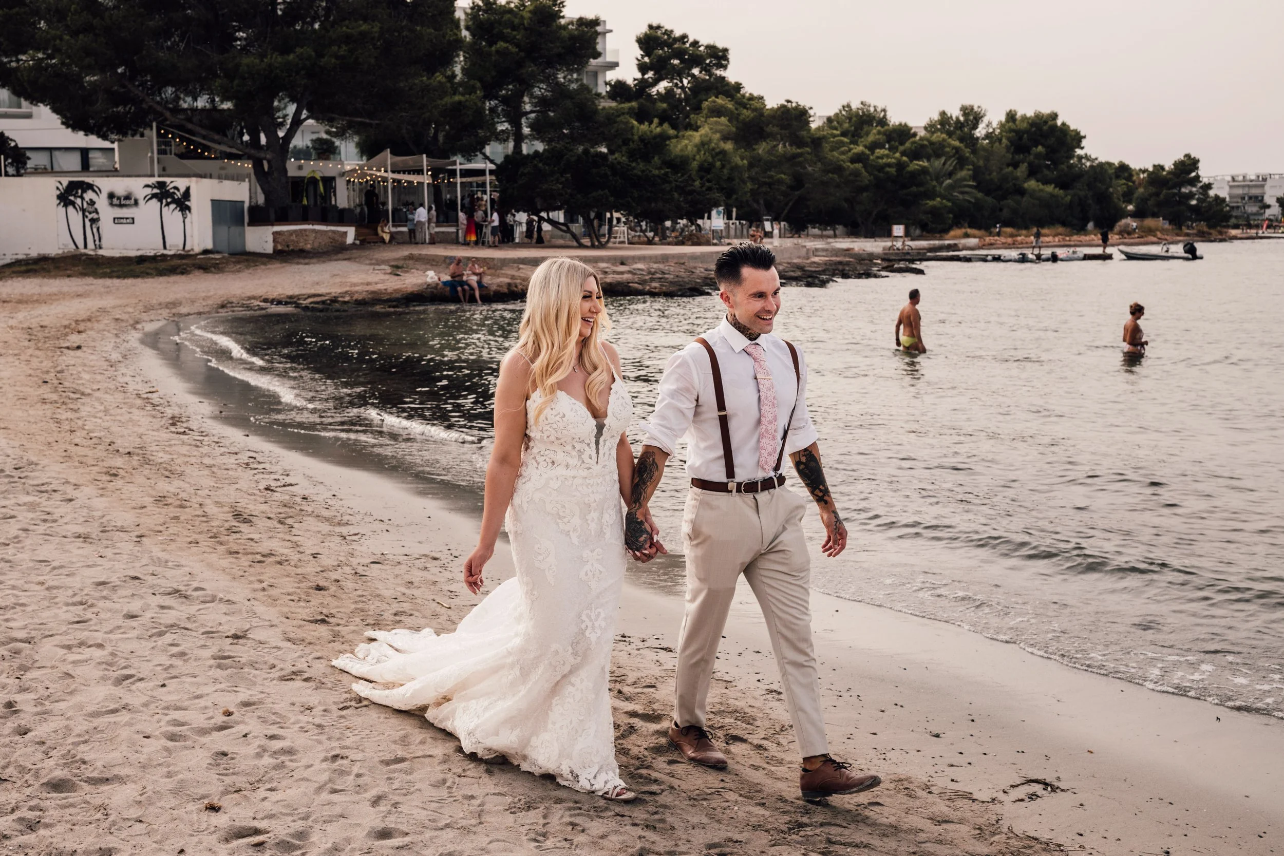 A newlywed couple walking hand in hand on the beach during sunset, with the bride in a white lace wedding dress and the groom in a white shirt, beige pants, and suspenders, smiling and enjoying their moment by the water.