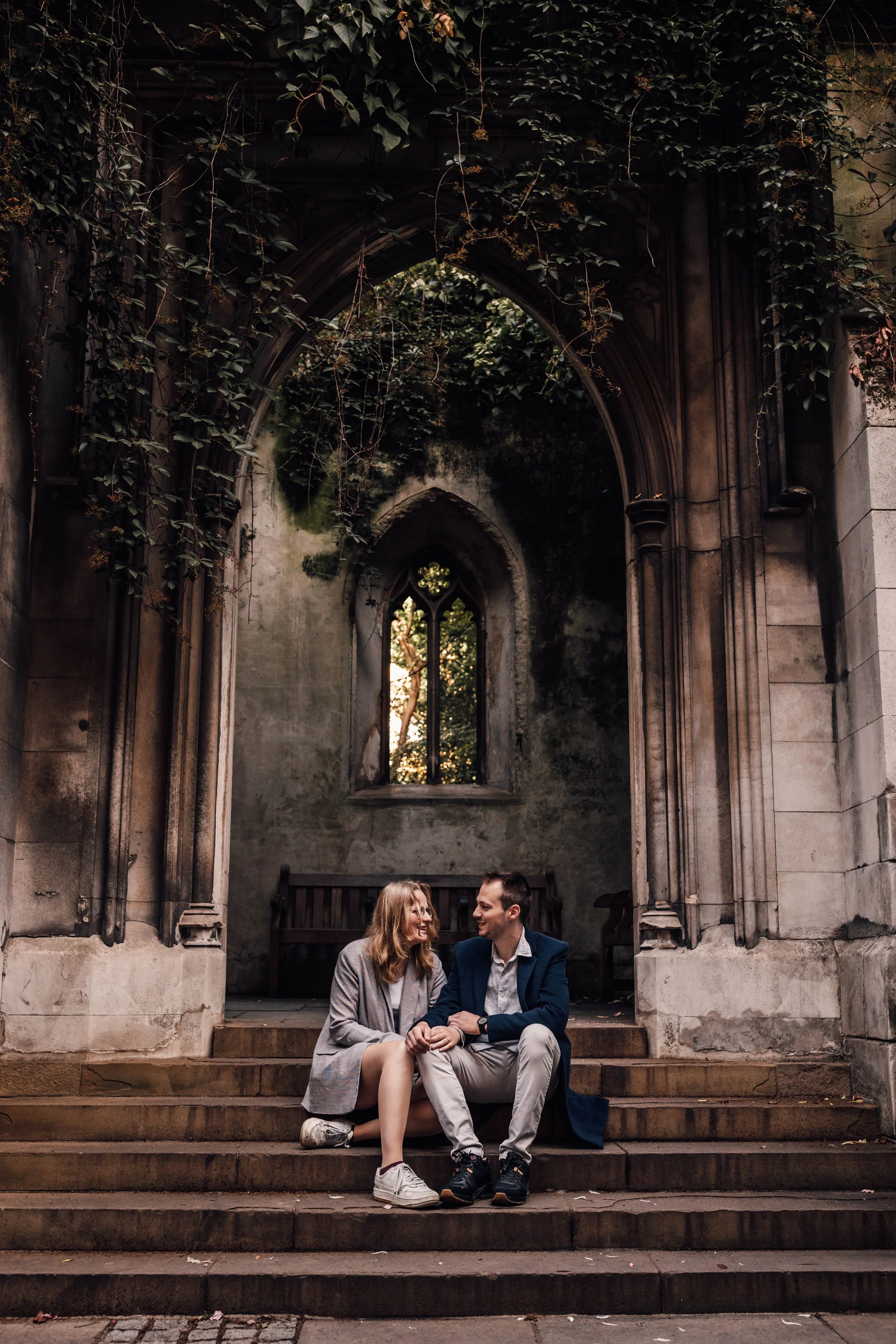 A young couple sits on stone steps outside an old, abandoned church with gothic-style arched window and overgrown ivy on the walls, sharing a moment of affection.